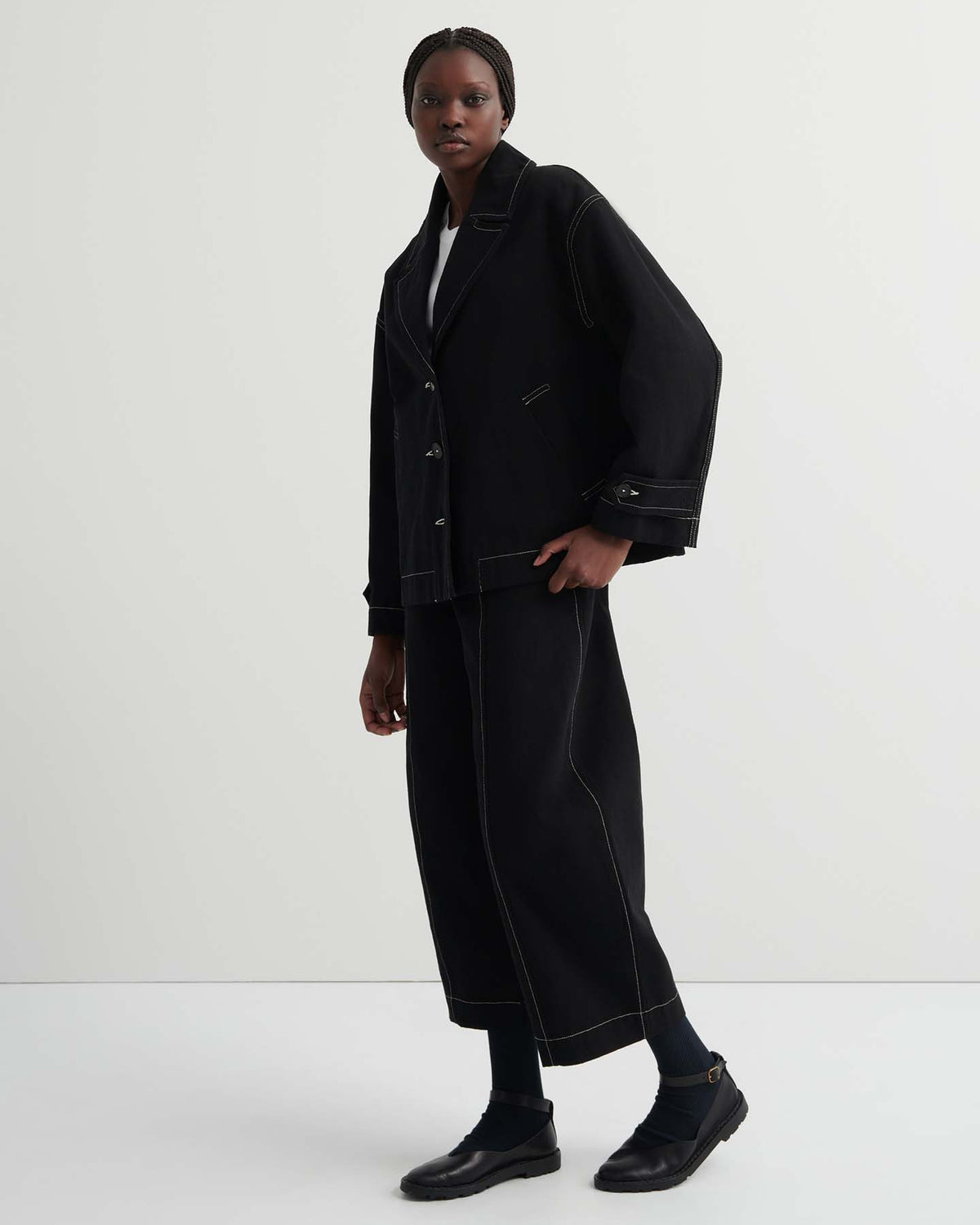 A person in the black Mural Jacket with a boxy, oversized fit, paired with matching wide-leg pants with contrast stitching, black shoes, and braided hair stands against a plain white background.