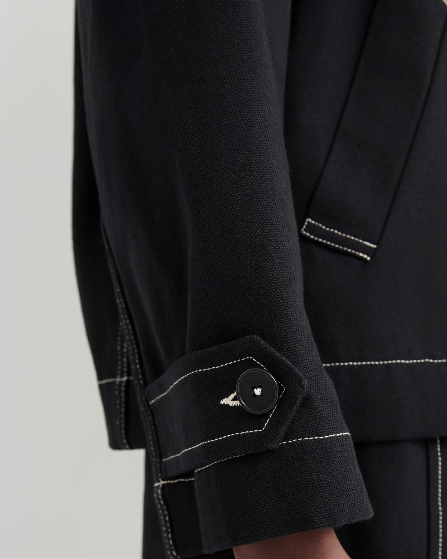 Someone stands against a plain light background wearing the oversized Mural Jacket – Black, featuring drop shoulder sleeves and white stitching, layered over a white shirt.