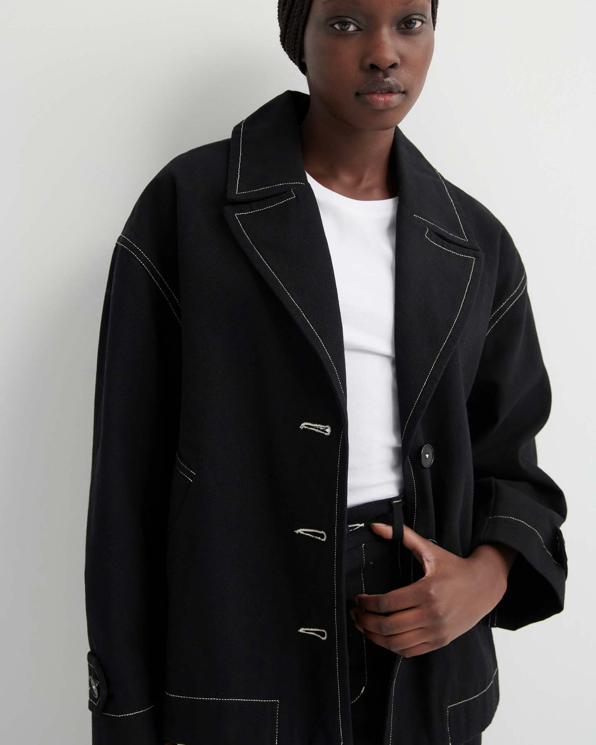 A person wearing the Mural Jacket – Black, an oversized fit with white stitching, over a white t-shirt and black pants stands against a plain light-colored wall.