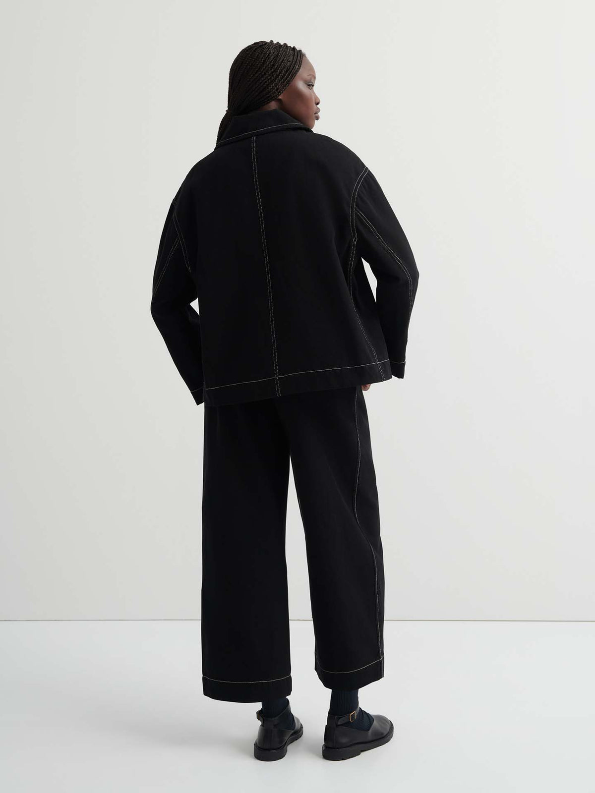 A person faces away, wearing the oversized Mural Jacket – Black with drop shoulder sleeves and wide-leg pants with visible white stitching, set against a plain light background.