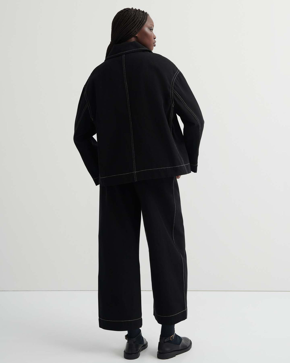 A person faces away, wearing the oversized Mural Jacket – Black with drop shoulder sleeves and wide-leg pants with visible white stitching, set against a plain light background.