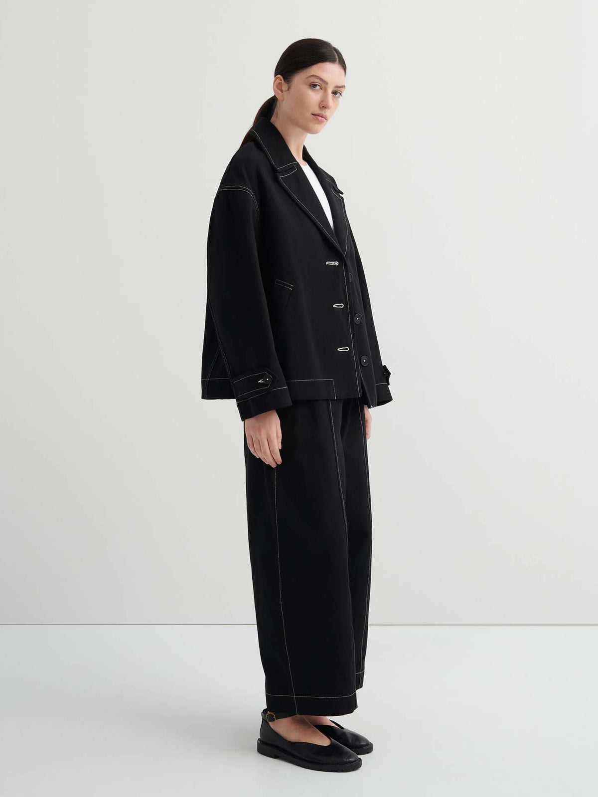 A person stands in a plain room wearing an oversized Mural Jacket – Black, matching wide-leg pants with white stitching, black shoes, and a white shirt.