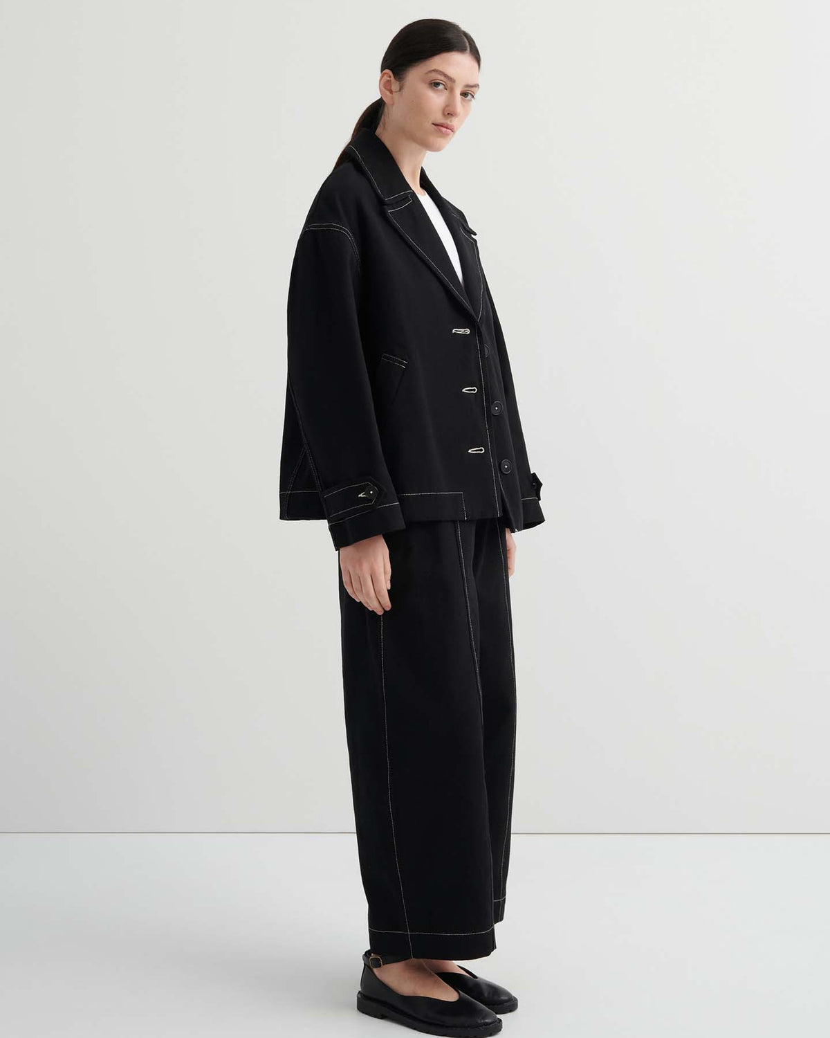 A person stands in a plain room wearing an oversized Mural Jacket – Black, matching wide-leg pants with white stitching, black shoes, and a white shirt.