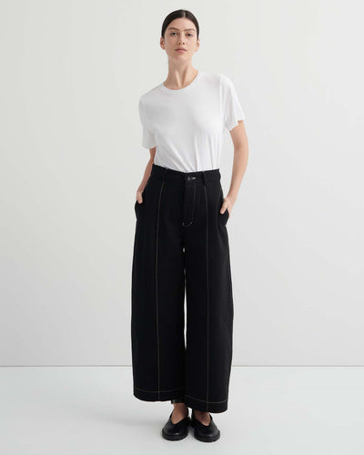 A person stands against a plain background, wearing a white t-shirt and the Mural Pant – Black with contrasting stitching and standard fit, paired with black shoes.