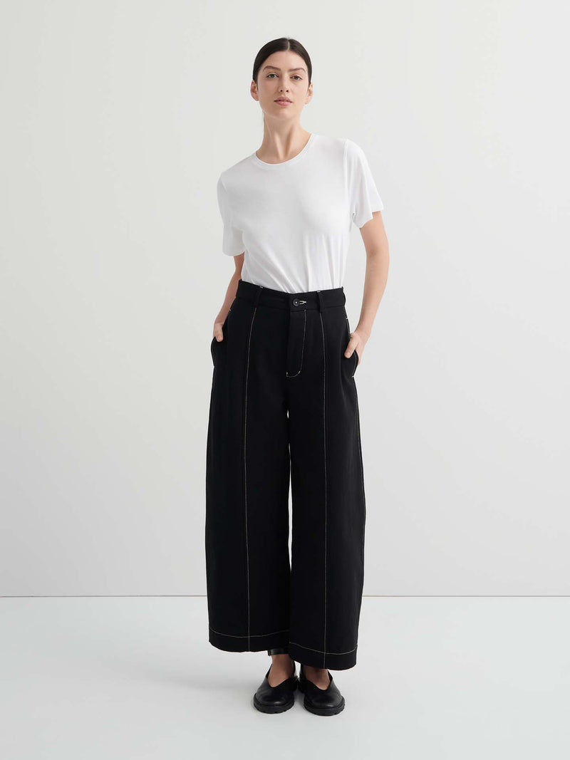 A person stands against a plain background, wearing a white t-shirt and the Mural Pant – Black with contrasting stitching and standard fit, paired with black shoes.