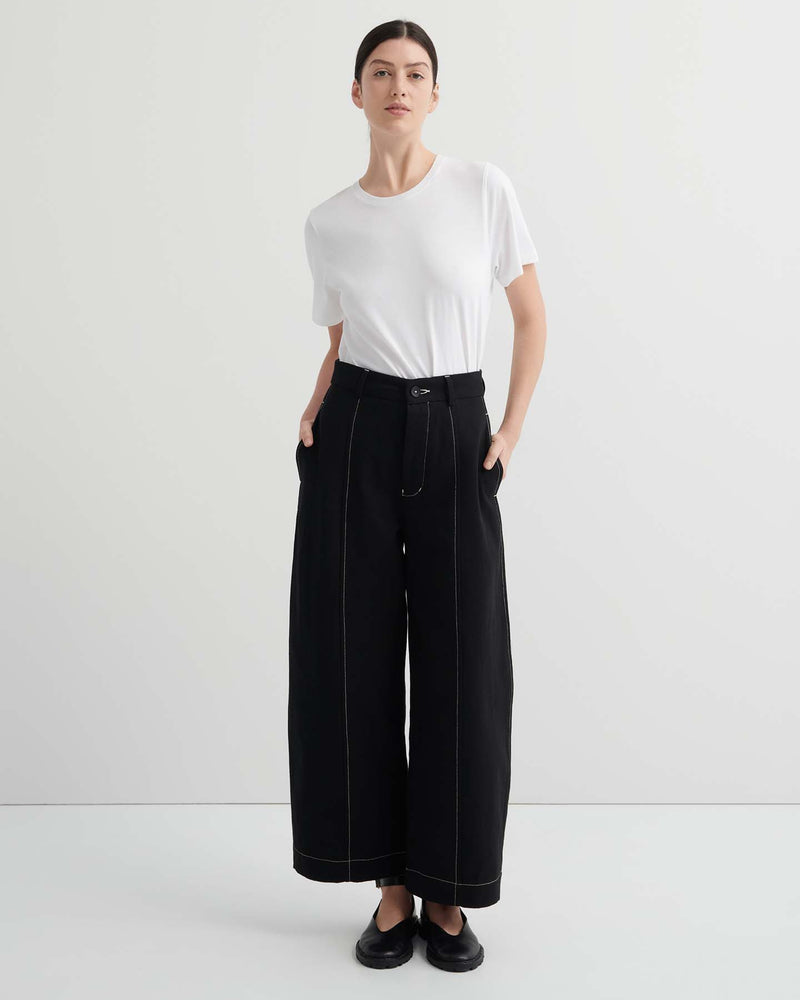 A person stands against a plain background, wearing a white t-shirt and the Mural Pant – Black with contrasting stitching and standard fit, paired with black shoes.
