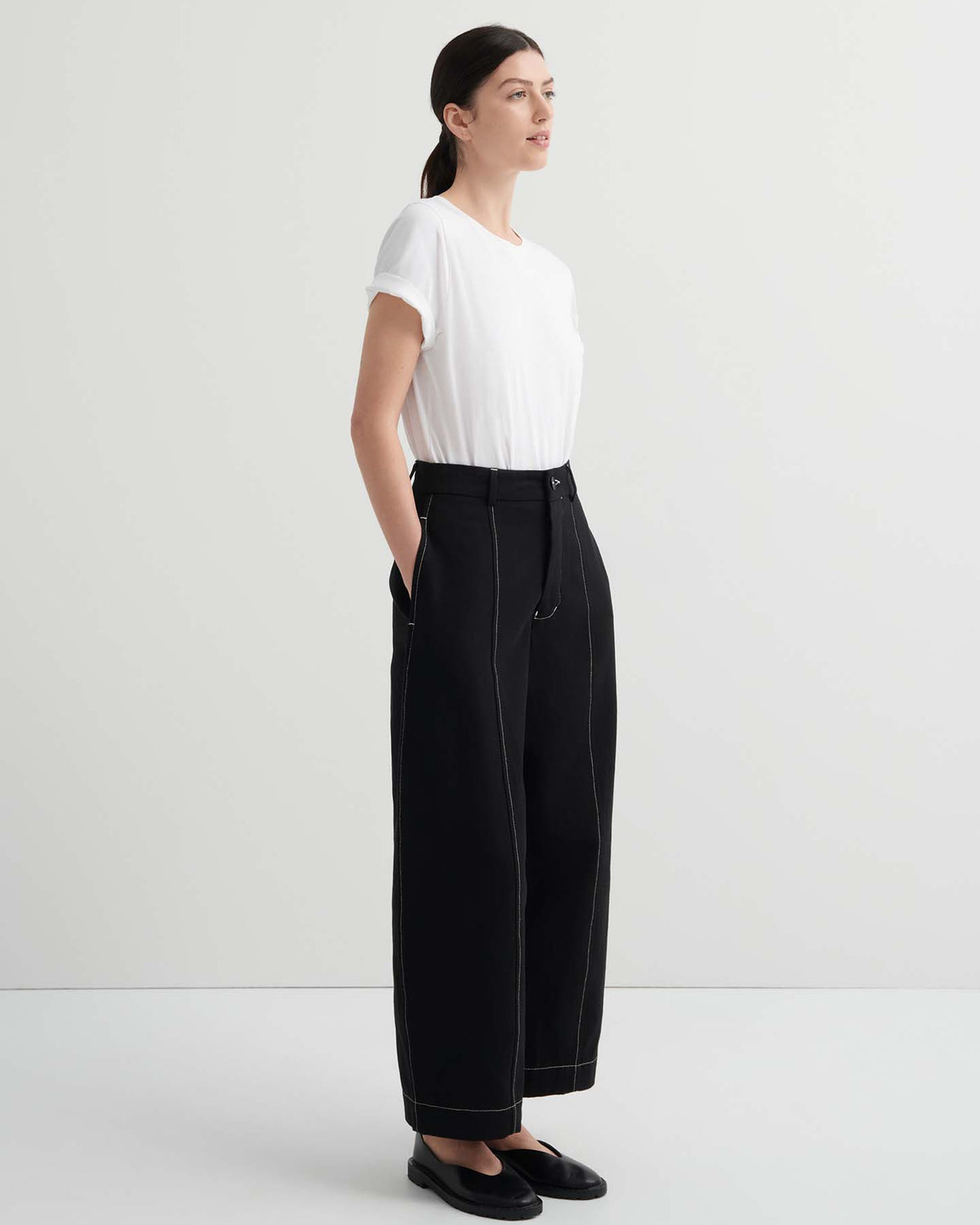 Woman stands against a plain background, wearing a white t-shirt, black flat shoes, and the Mural Pant – Black in a high-rise, standard fit with visible seams.