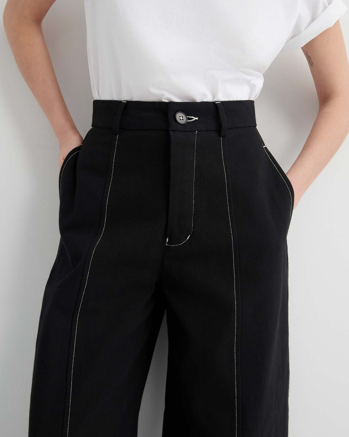 Model in Mural Pant – Black, featuring white contrast stitching and a white short-sleeve shirt, hands in pockets. Shown from shoulders to mid-thigh. The standard fit combines comfort and style, ideal for everyday wear.