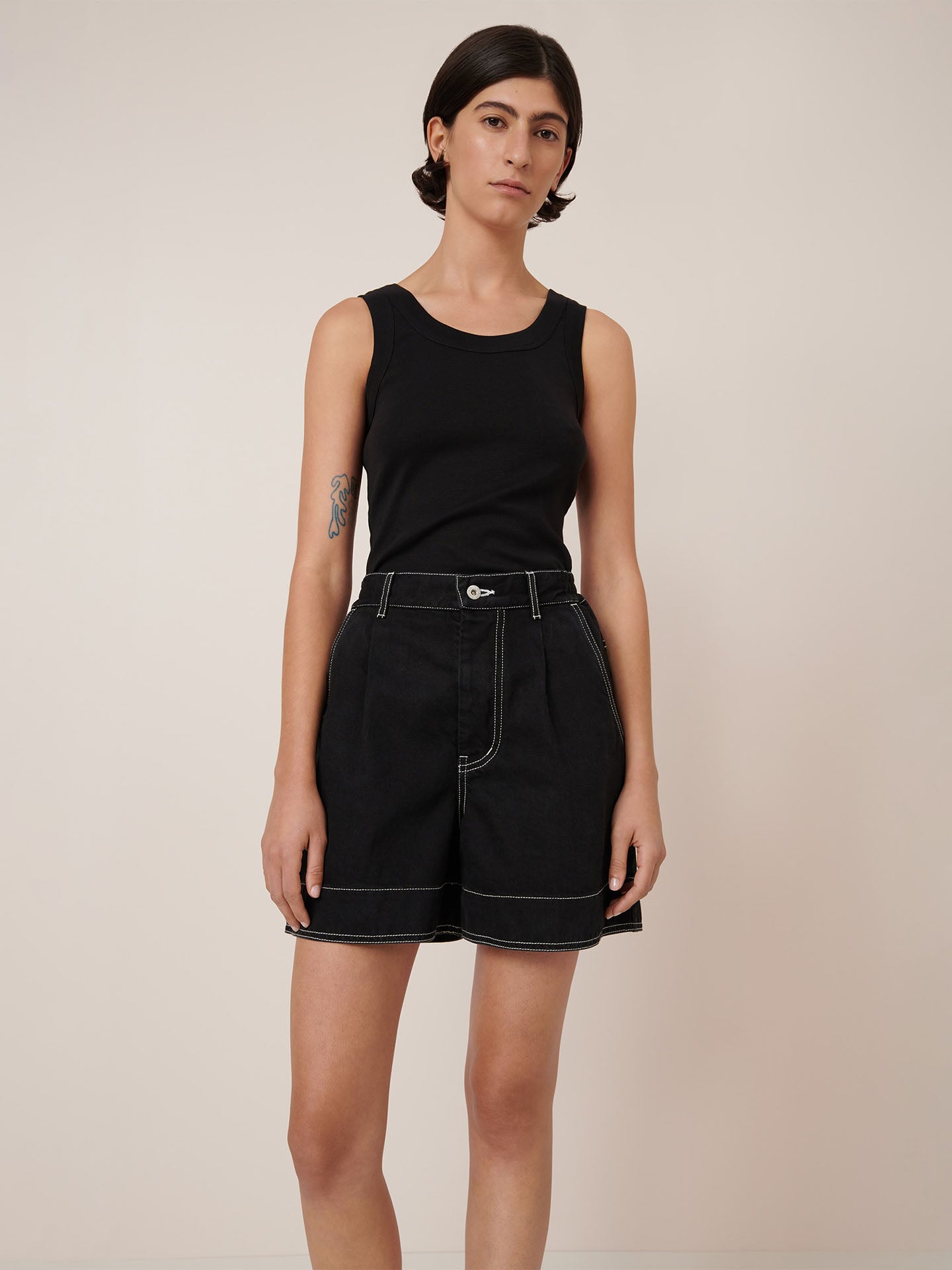 A person with short dark hair wears the Kowtow Outline Shorts in black, featuring white stitching and an elastic waistband, paired with a black sleeveless top, standing against a plain light background.