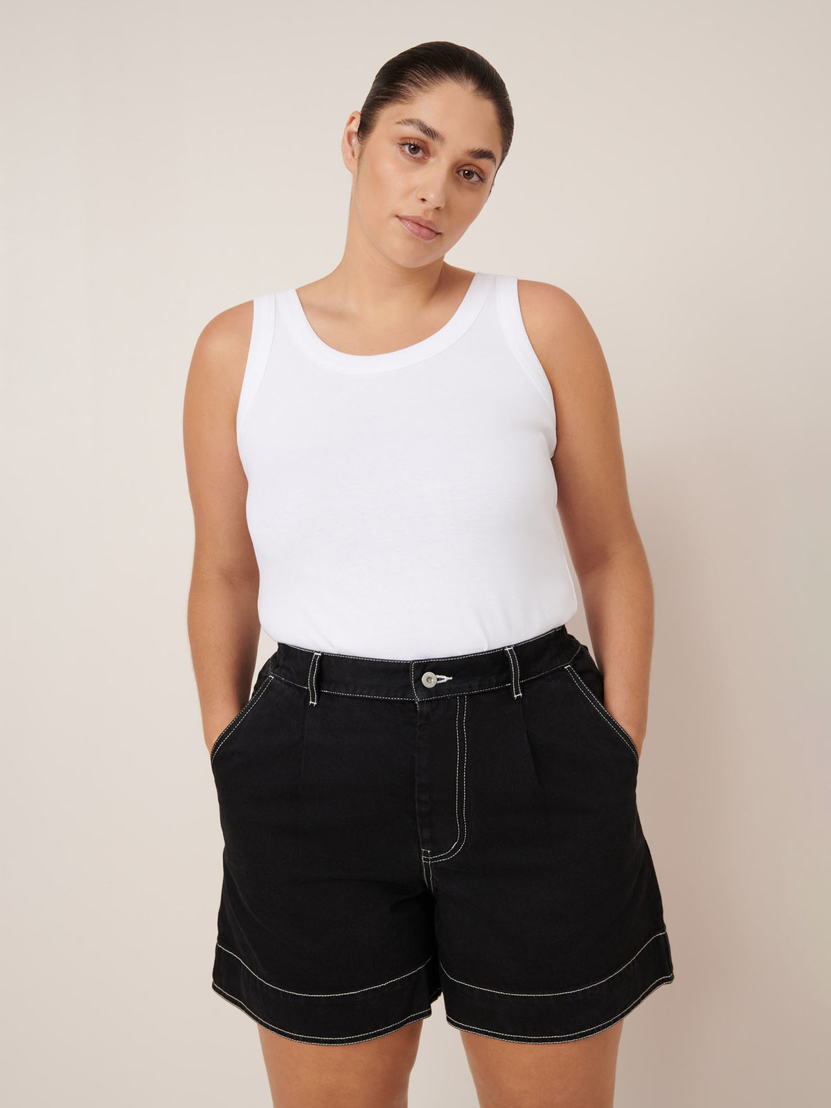 A person wears Kowtow&#39;s Outline Shorts in black, styled with a white sleeveless top, standing hands-in-pockets against a plain light background.