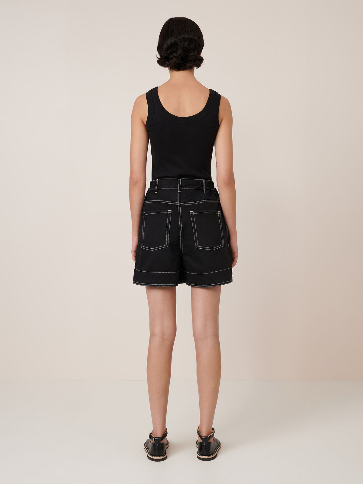 A person stands facing away from the camera, wearing Kowtow&#39;s Outline Shorts in black with an elastic waistband and black sandals, set against a plain background.