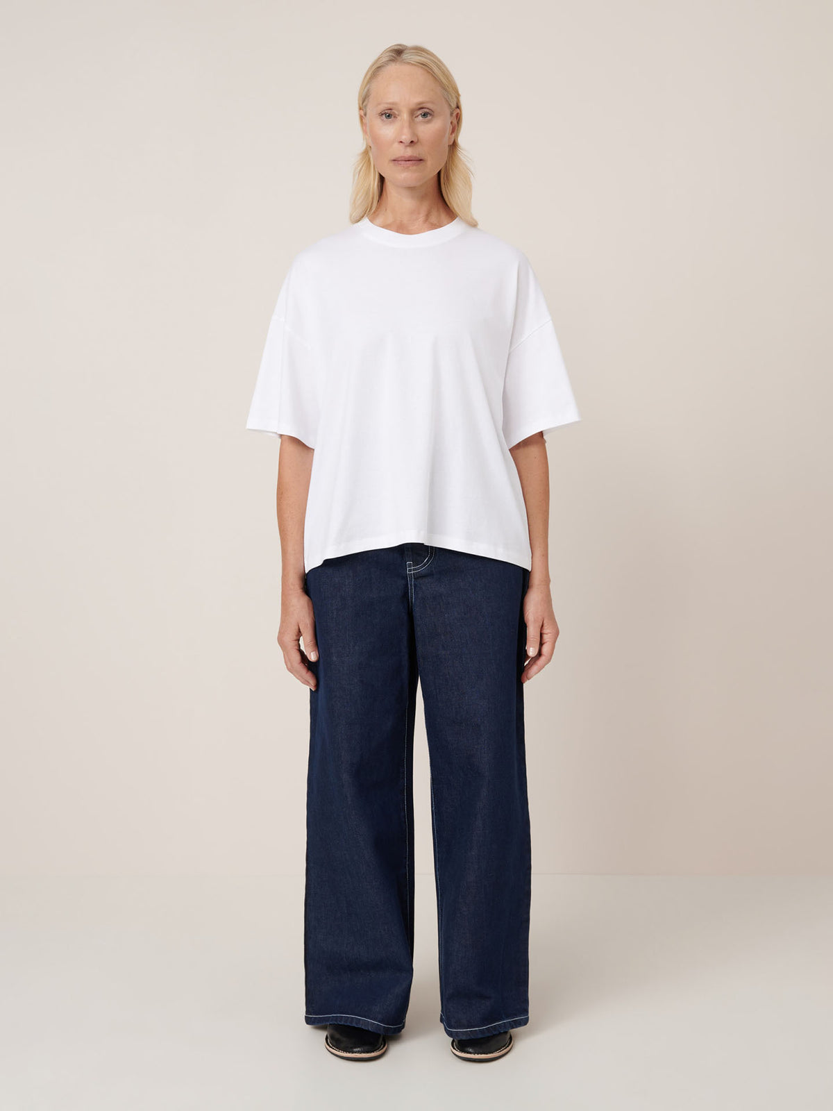 A woman faces forward against a beige background, wearing the Kowtow Oversized Boxy Tee in white with drop shoulders, paired with wide-leg dark blue jeans and black shoes.
