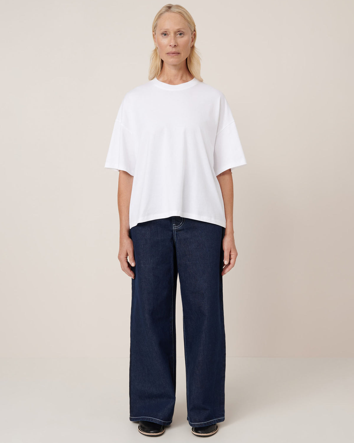A woman faces forward against a beige background, wearing the Kowtow Oversized Boxy Tee in white with drop shoulders, paired with wide-leg dark blue jeans and black shoes.