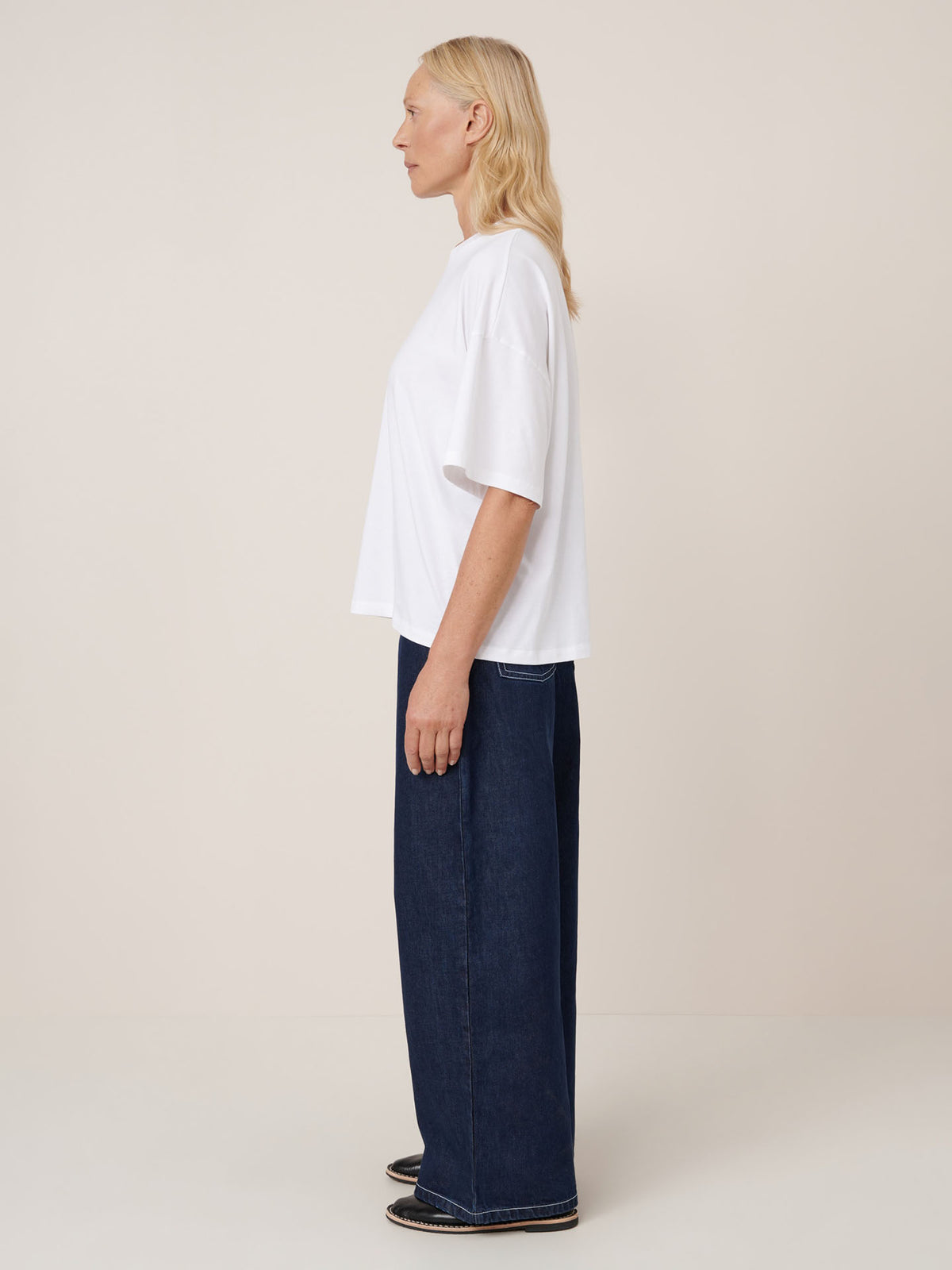 A person stands in profile against a plain background, wearing the Kowtow Oversized Boxy Tee in white, paired with wide-leg dark blue jeans and black shoes.