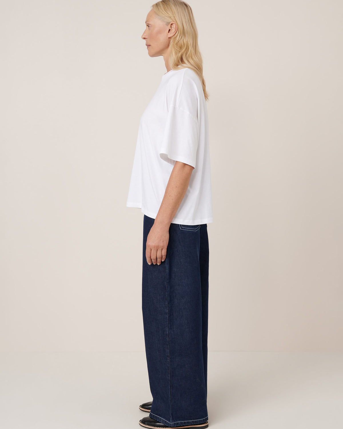 A person stands in profile against a plain background, wearing the Kowtow Oversized Boxy Tee in white, paired with wide-leg dark blue jeans and black shoes.