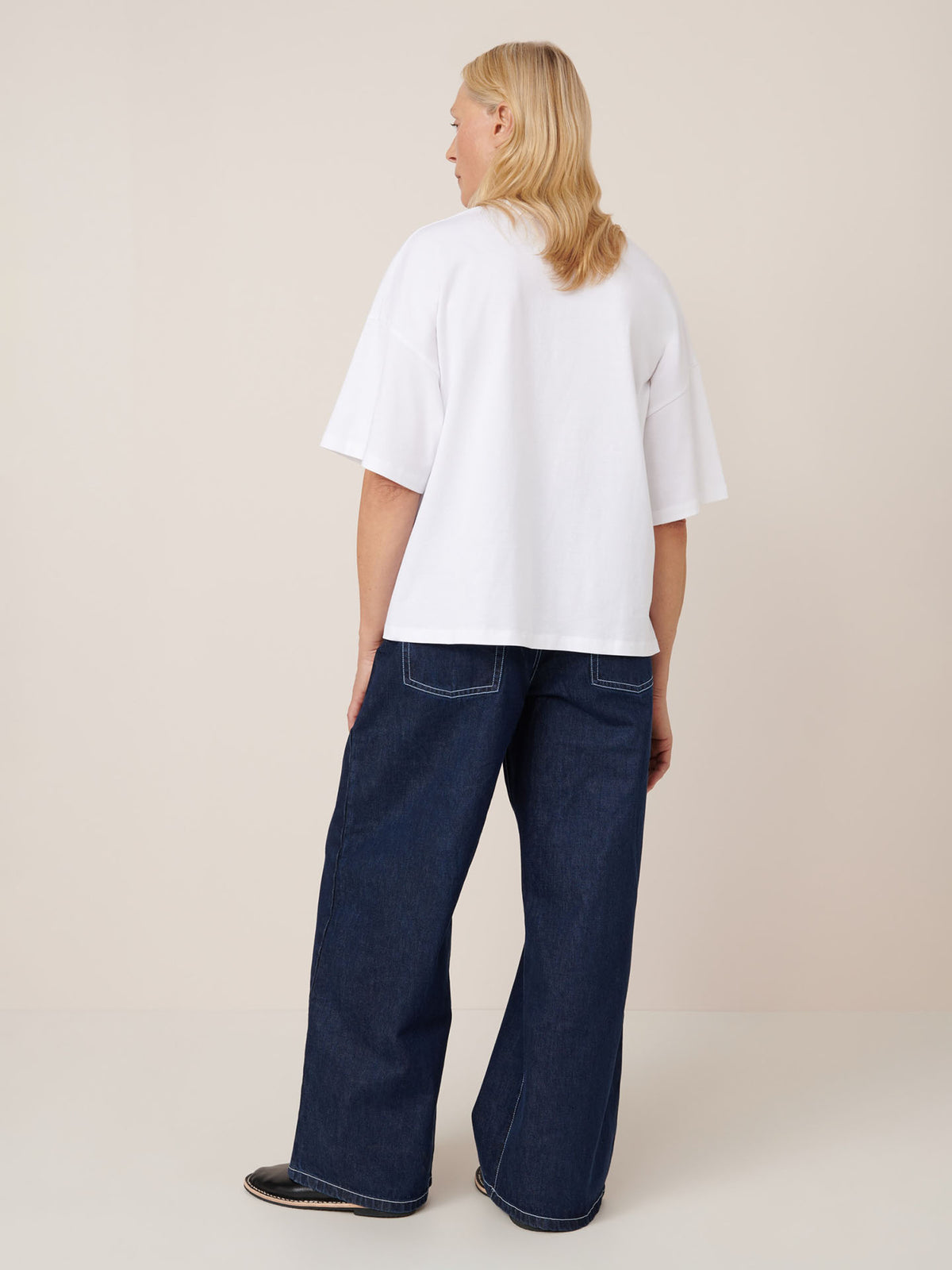 A person faces away, wearing the Kowtow Oversized Boxy Tee in white with drop shoulder sleeves and wide-leg dark blue jeans, set against a plain light background.
