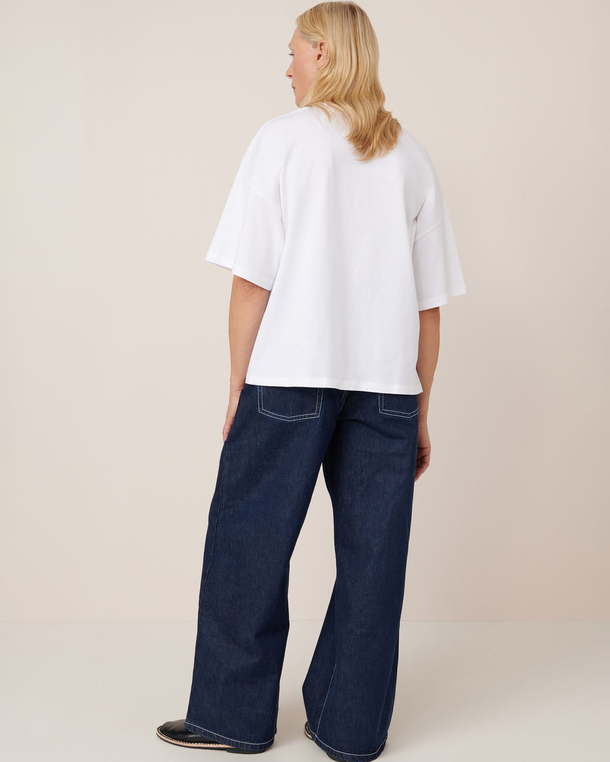 A person faces away, wearing the Kowtow Oversized Boxy Tee in white with drop shoulder sleeves and wide-leg dark blue jeans, set against a plain light background.