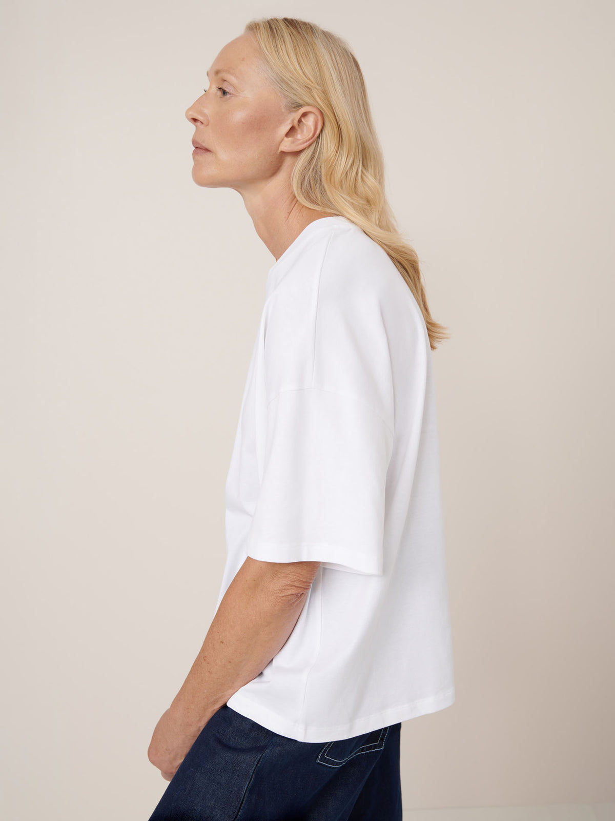 A person with long blond hair is shown in side profile against a plain background, wearing dark jeans and the Kowtow Oversized Boxy Tee in white with drop shoulder sleeves.