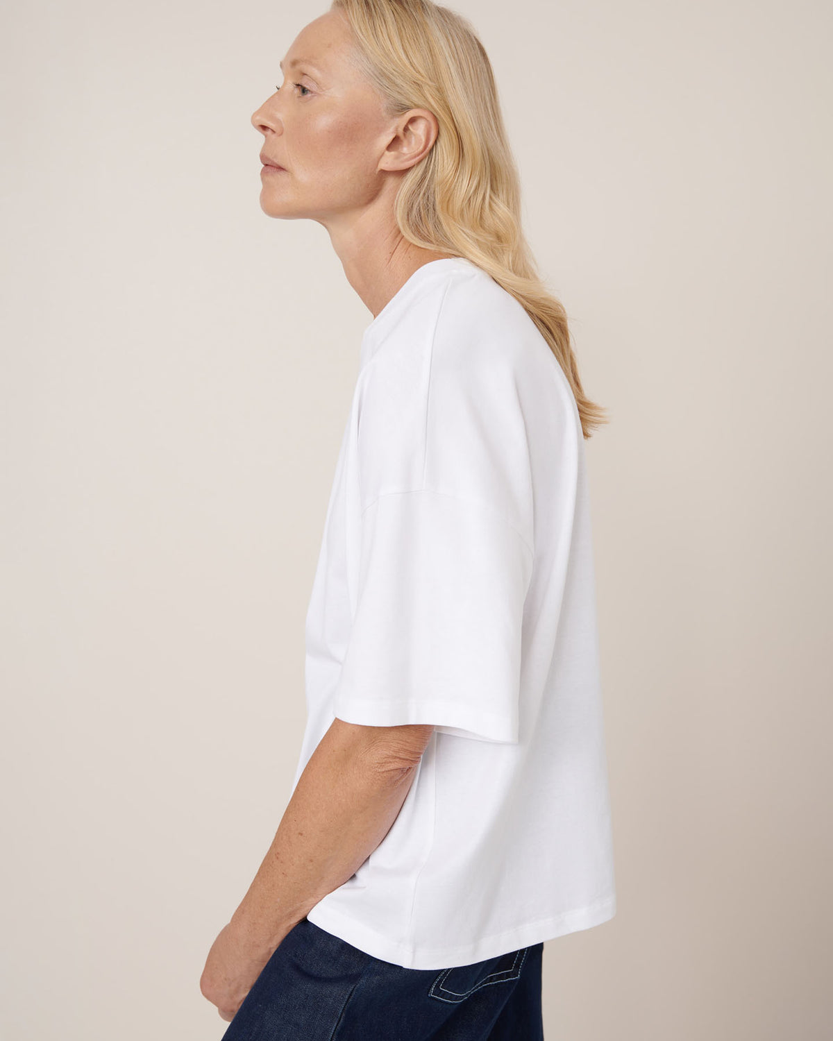 A person with long blond hair is shown in side profile against a plain background, wearing dark jeans and the Kowtow Oversized Boxy Tee in white with drop shoulder sleeves.
