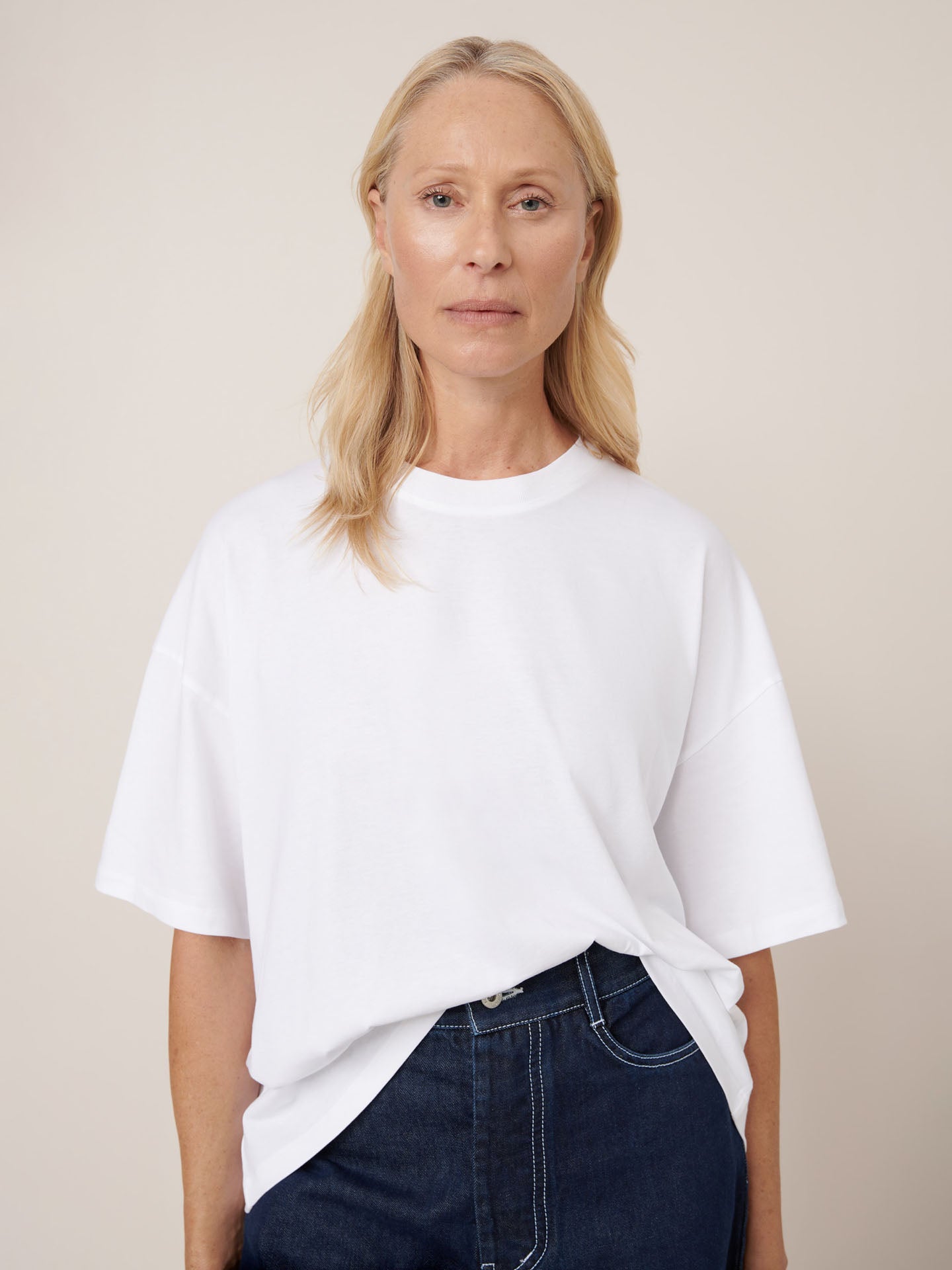 A woman with long blonde hair wears the Kowtow Oversized Boxy Tee in white, paired with dark jeans, standing against a neutral background and looking directly at the camera.