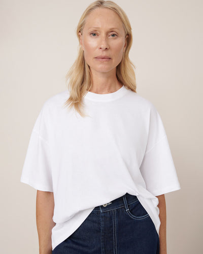 A woman with long blonde hair wears the Kowtow Oversized Boxy Tee in white, paired with dark jeans, standing against a neutral background and looking directly at the camera.