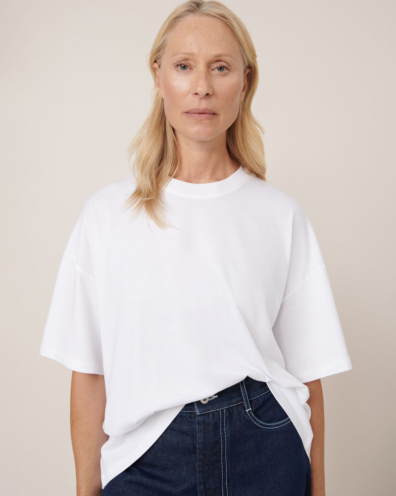 A woman with long blonde hair wears the Kowtow Oversized Boxy Tee in white, paired with dark jeans, standing against a neutral background and looking directly at the camera.