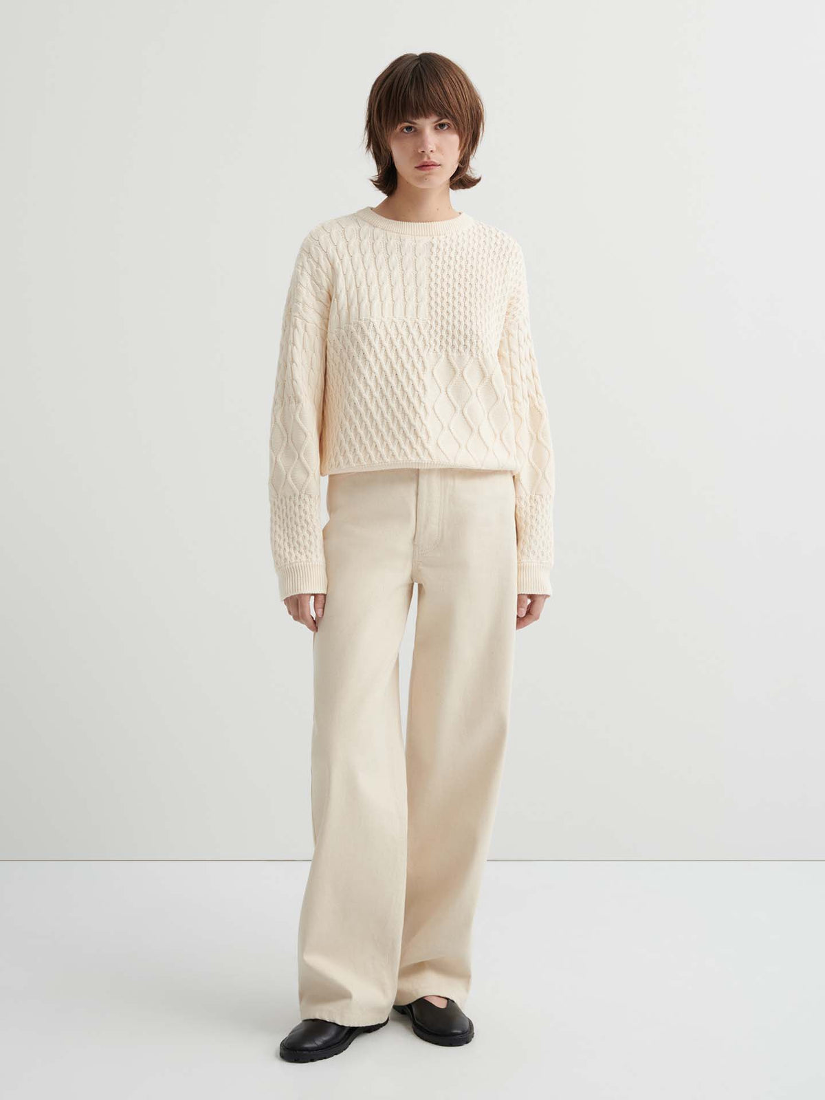 Person standing against a plain background, wearing the Patchwork Jumper – Chalk, an oversized cream textured sweater with a boxy silhouette, paired with wide-leg cream pants and black shoes.