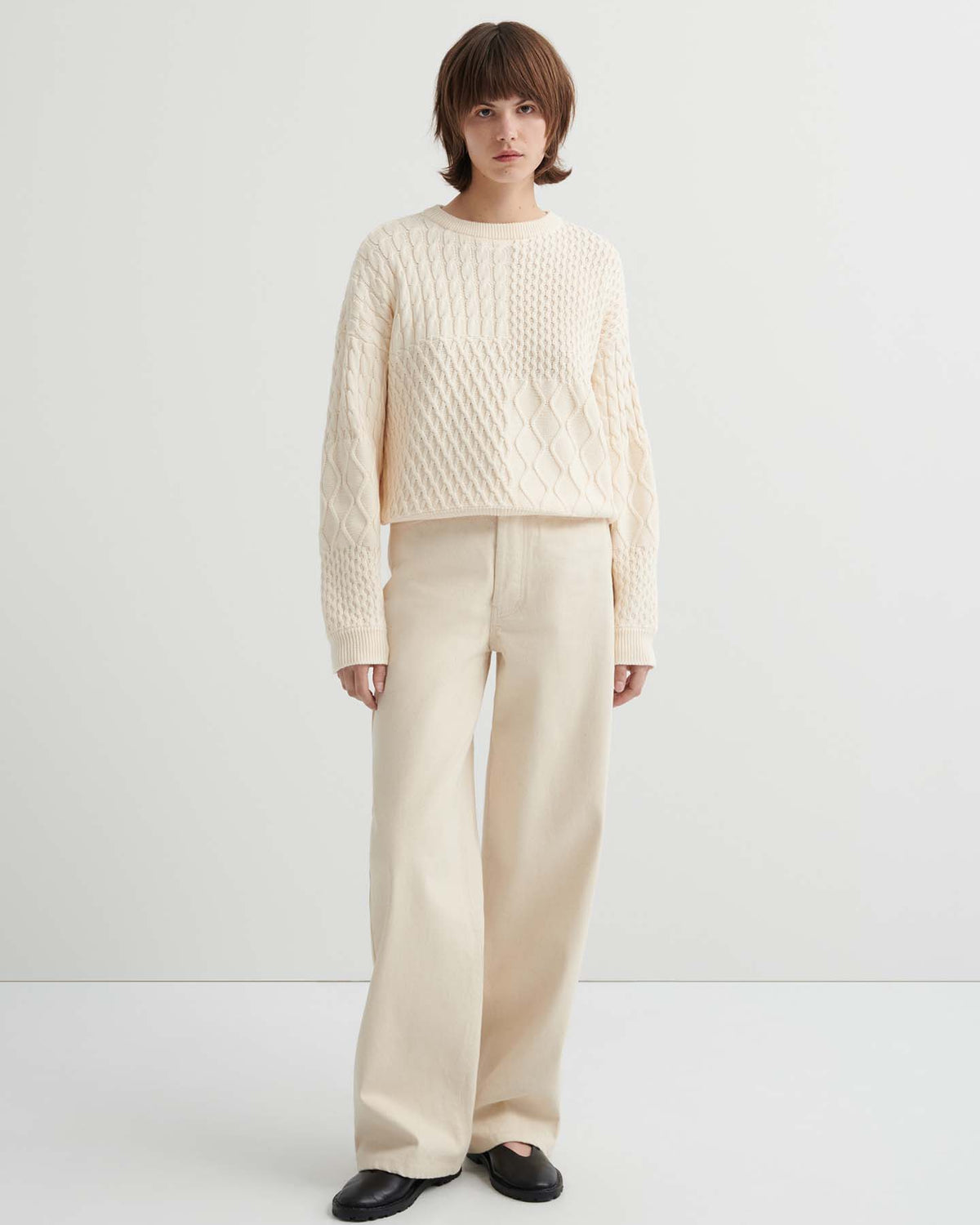 Person standing against a plain background, wearing the Patchwork Jumper – Chalk, an oversized cream textured sweater with a boxy silhouette, paired with wide-leg cream pants and black shoes.
