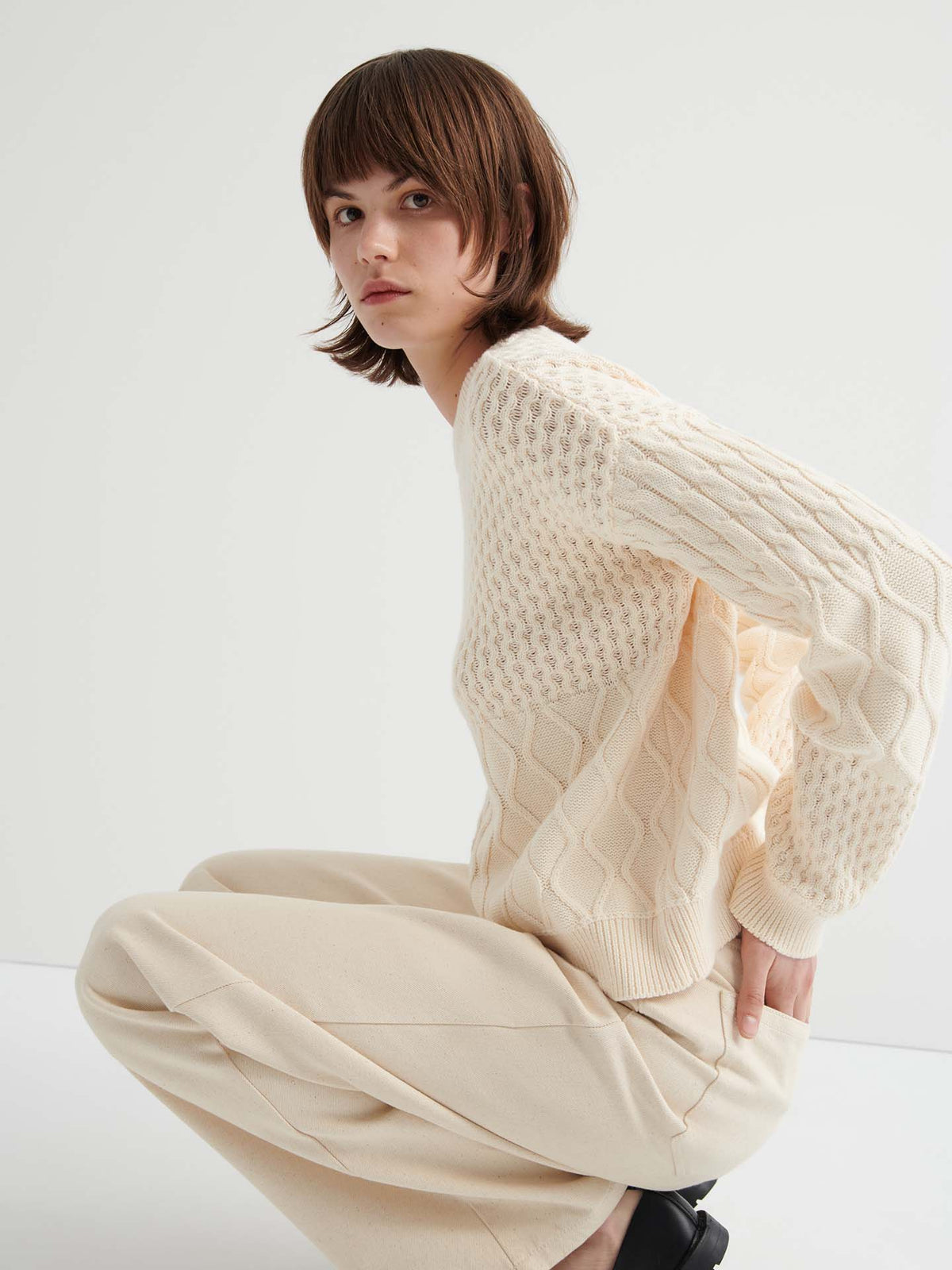 A person with short brown hair wears the Patchwork Jumper – Chalk in an oversized fit with beige pants, crouching against a plain white background.