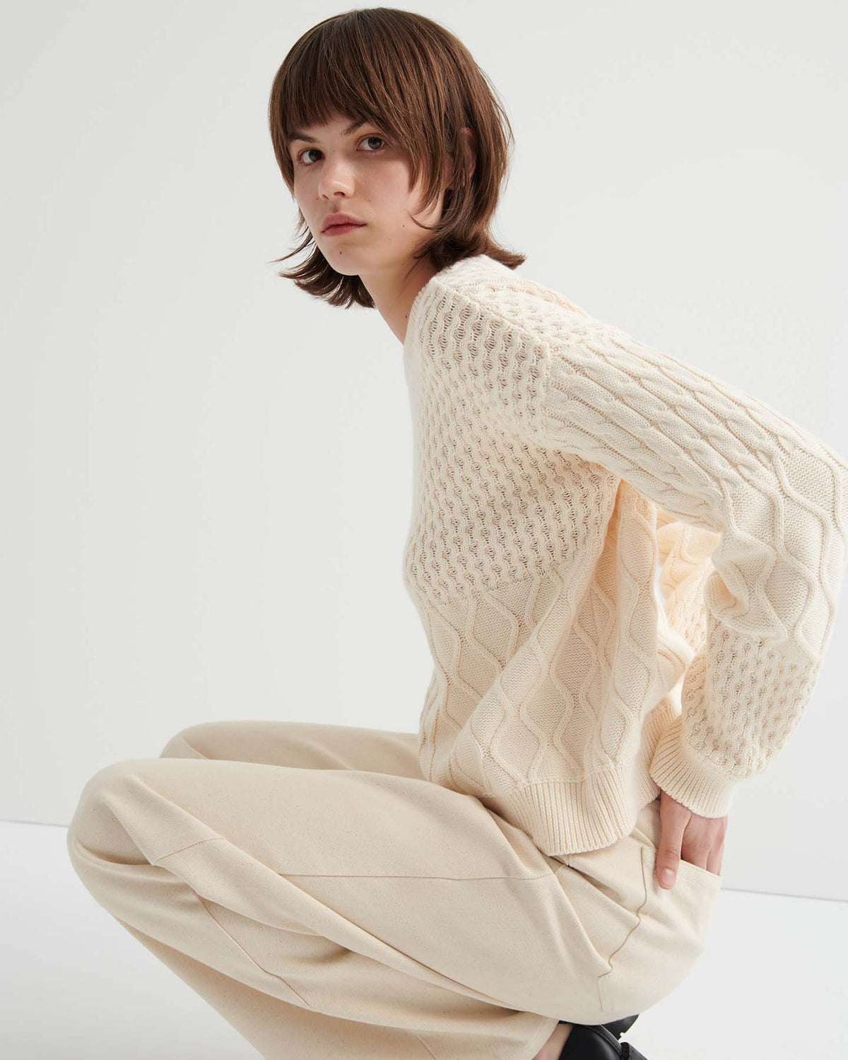 A person with short brown hair wears the Patchwork Jumper – Chalk in an oversized fit with beige pants, crouching against a plain white background.