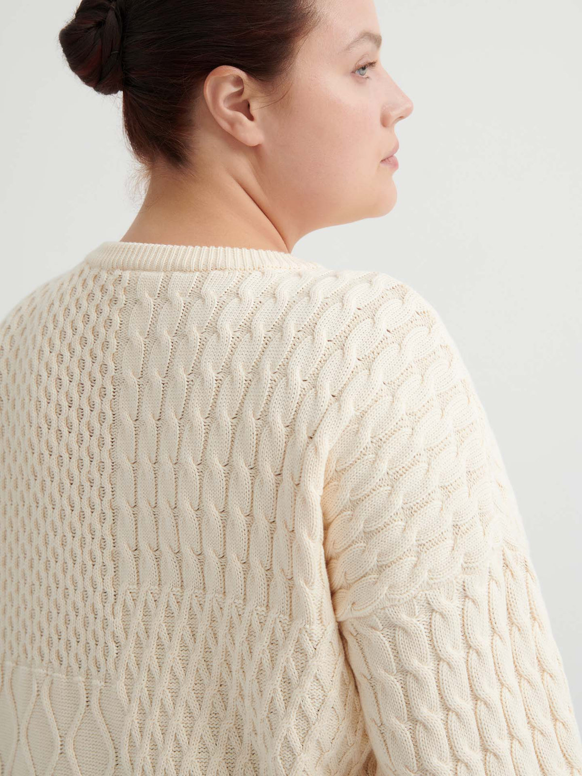 A woman with dark hair in a bun models the Patchwork Jumper – Chalk, a cream-hued textured knit with an oversized fit, shown from the back and in profile against a plain light background.