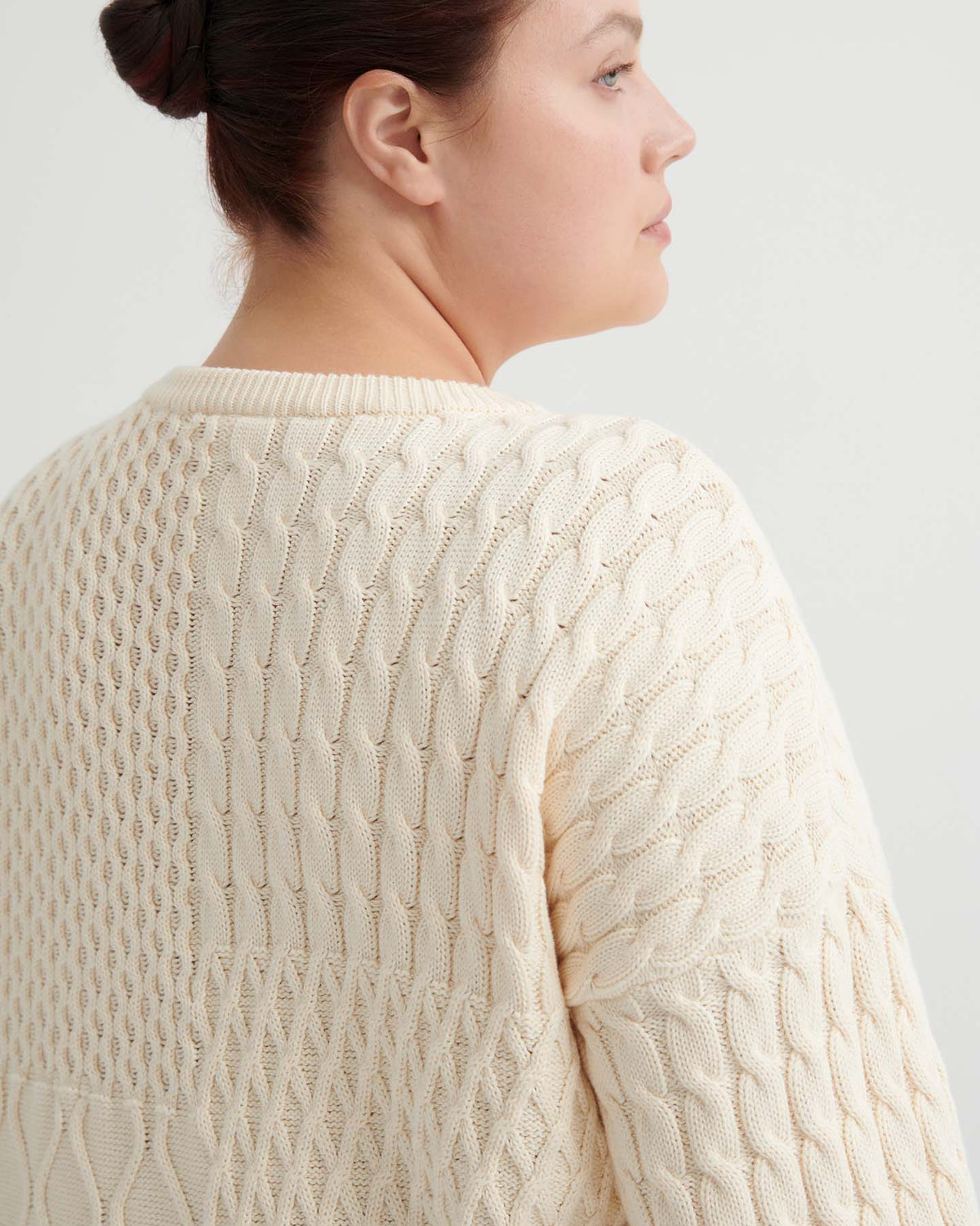 A woman with dark hair in a bun models the Patchwork Jumper – Chalk, a cream-hued textured knit with an oversized fit, shown from the back and in profile against a plain light background.