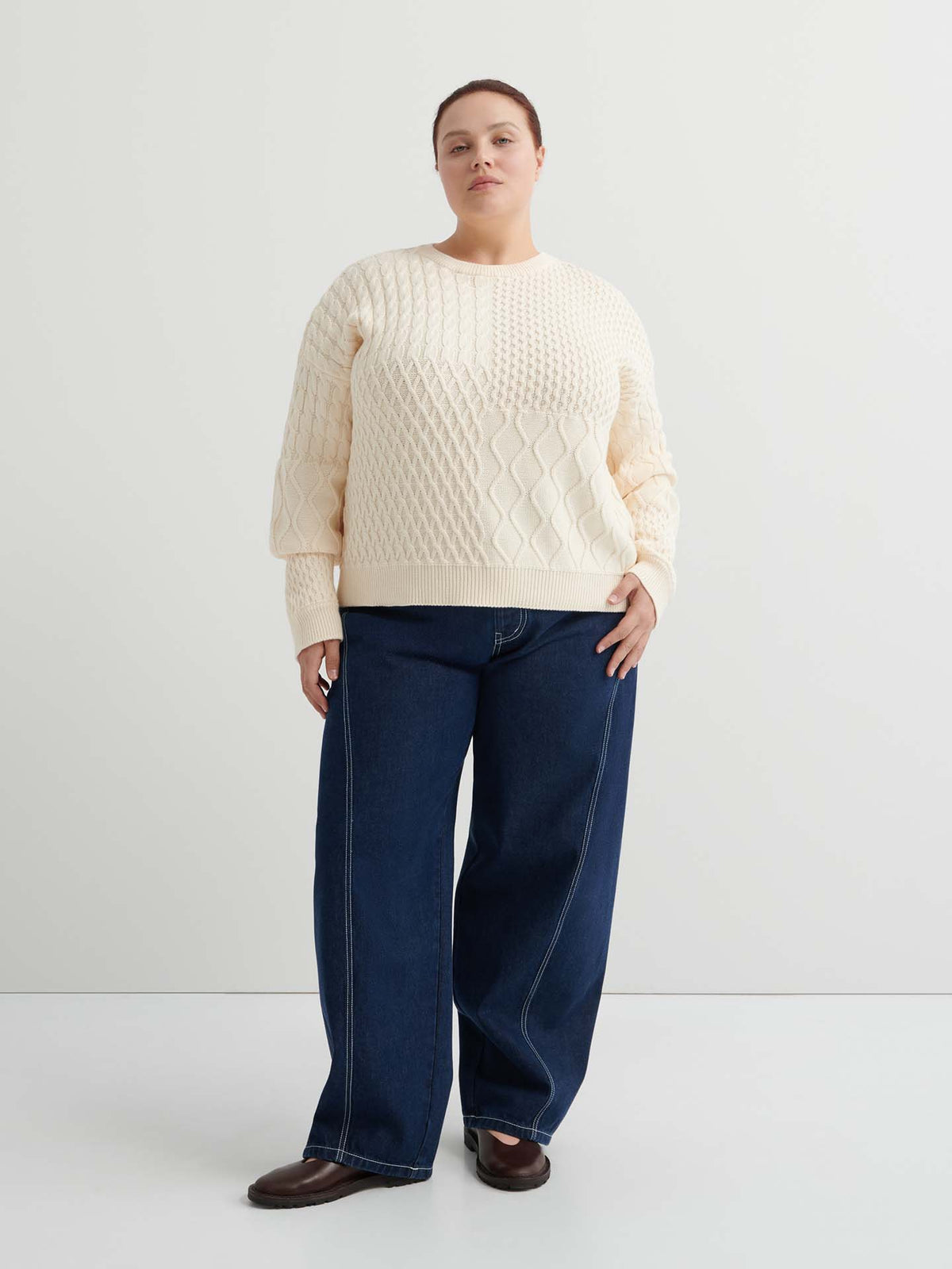 A person stands against a plain white background, wearing the Patchwork Jumper – Chalk with a boxy silhouette, paired with wide-leg dark blue jeans and brown shoes.
