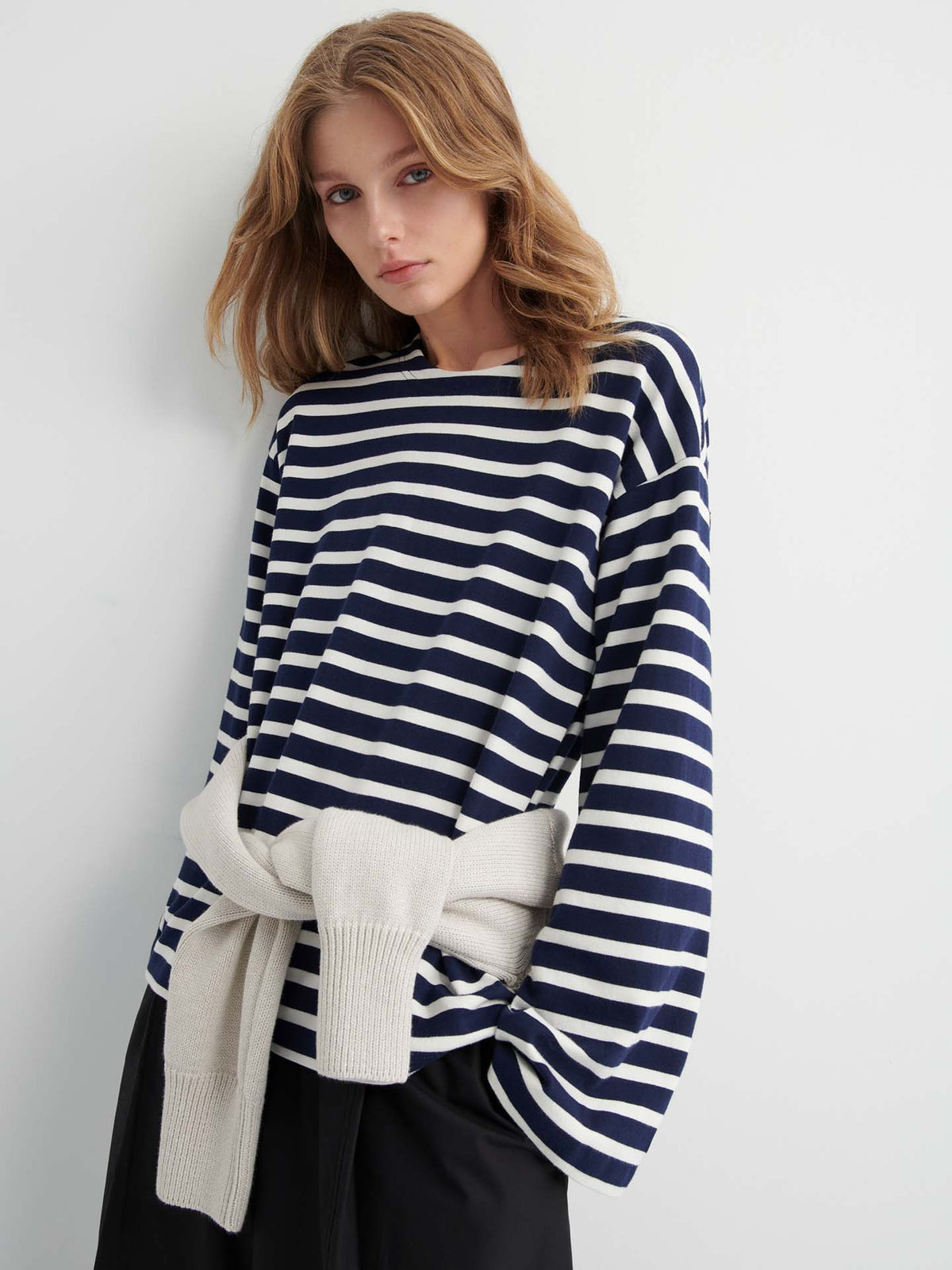 A woman stands against a plain wall wearing the Pierre Tee – Uniform Stripe, a navy and white striped long-sleeve top with a relaxed fit, paired with black pants and a light sweater tied around her waist.