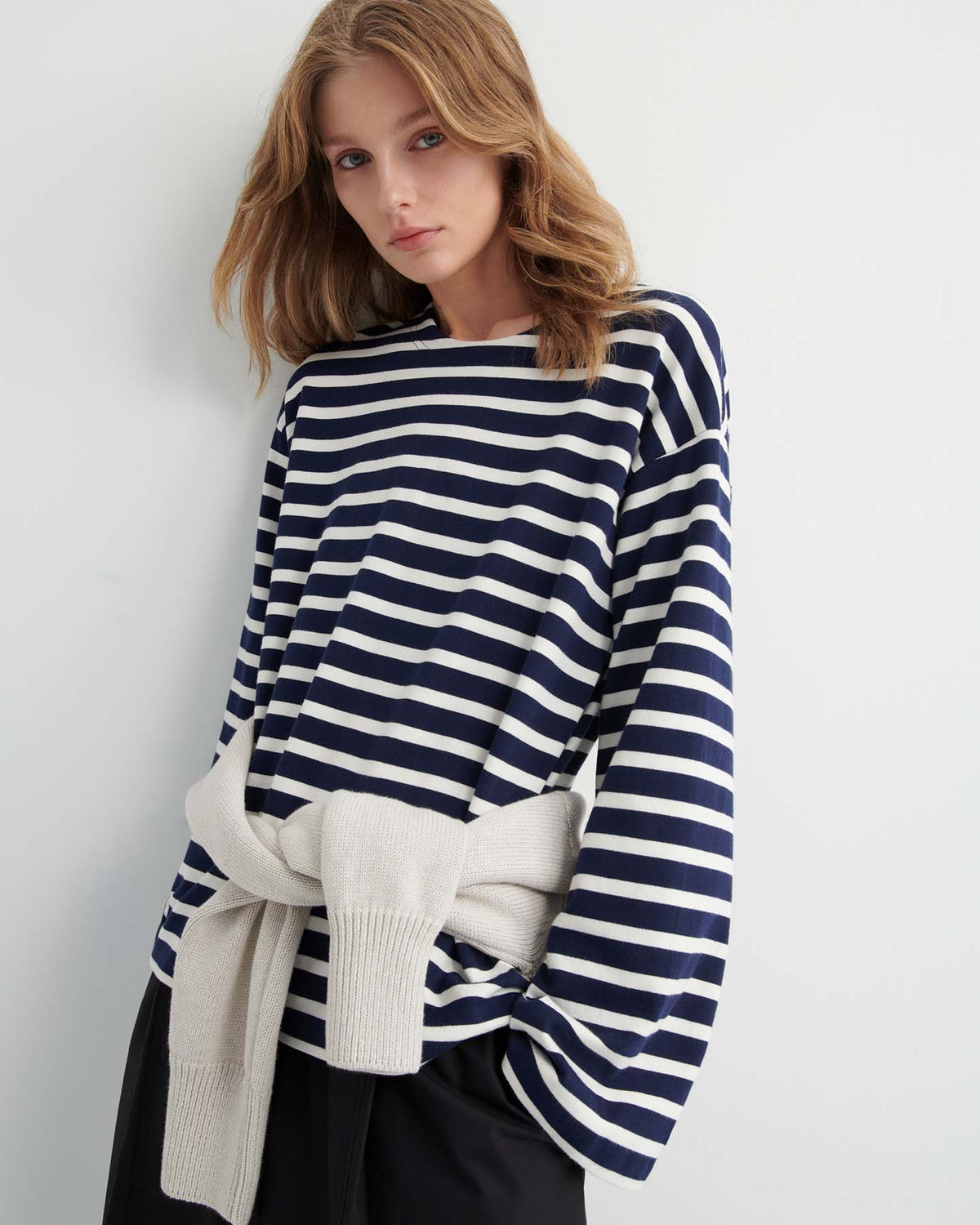 A woman stands against a plain wall wearing the Pierre Tee – Uniform Stripe, a navy and white striped long-sleeve top with a relaxed fit, paired with black pants and a light sweater tied around her waist.