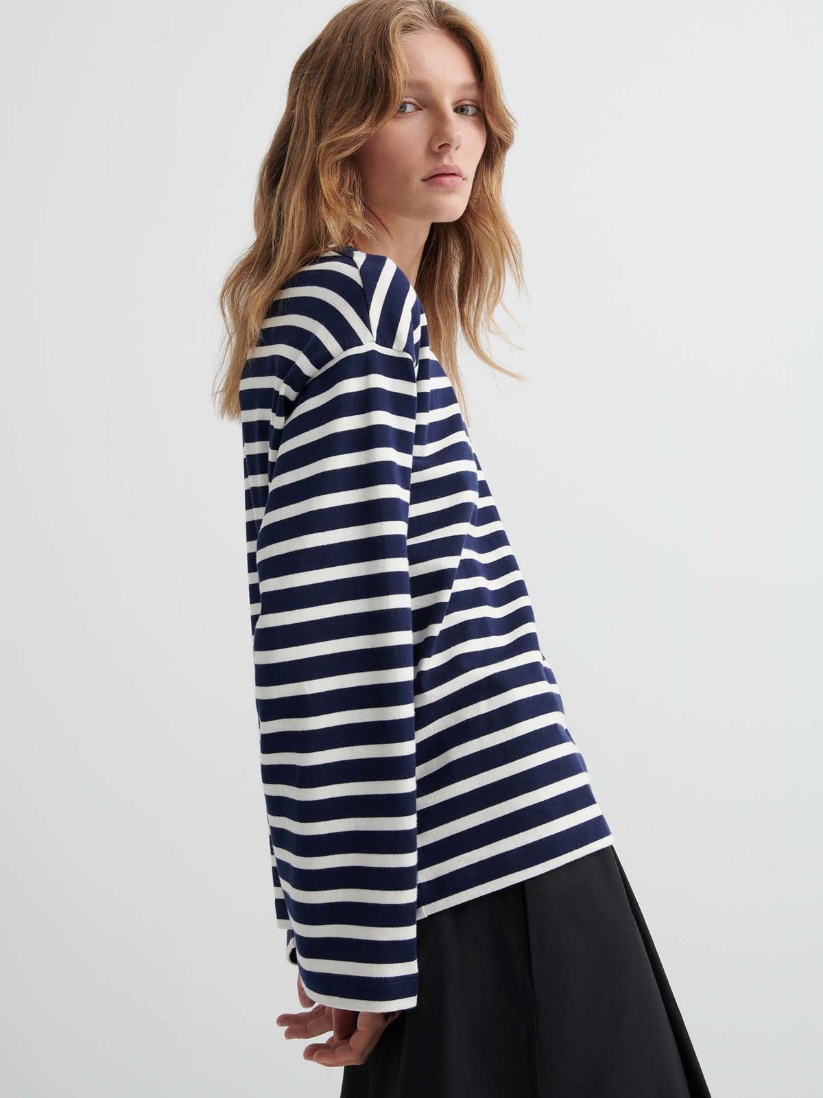 A person with long, wavy hair stands sideways against a plain background, wearing the Pierre Tee – Uniform Stripe in navy and white with black pants.