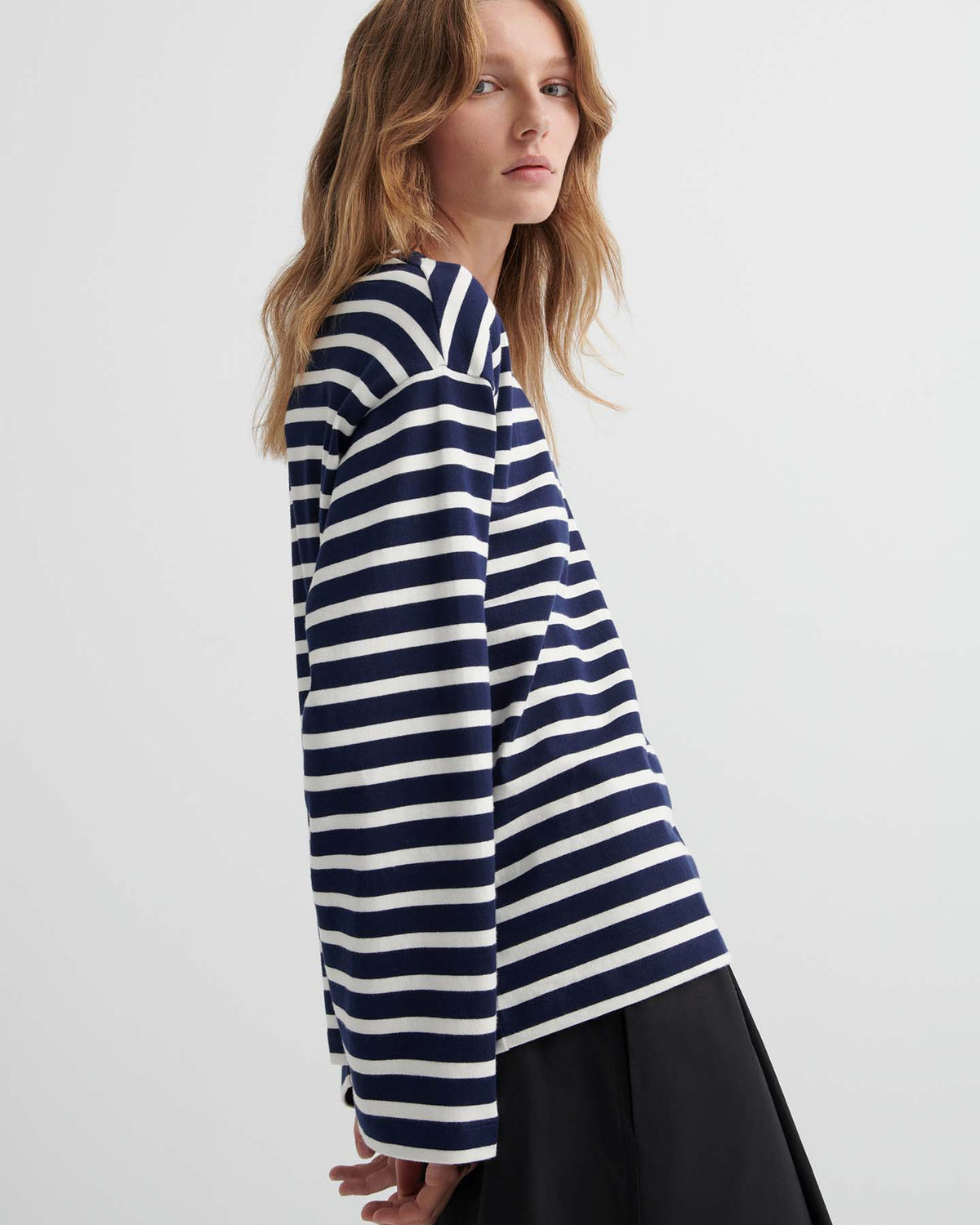 A person with long, wavy hair stands sideways against a plain background, wearing the Pierre Tee – Uniform Stripe in navy and white with black pants.