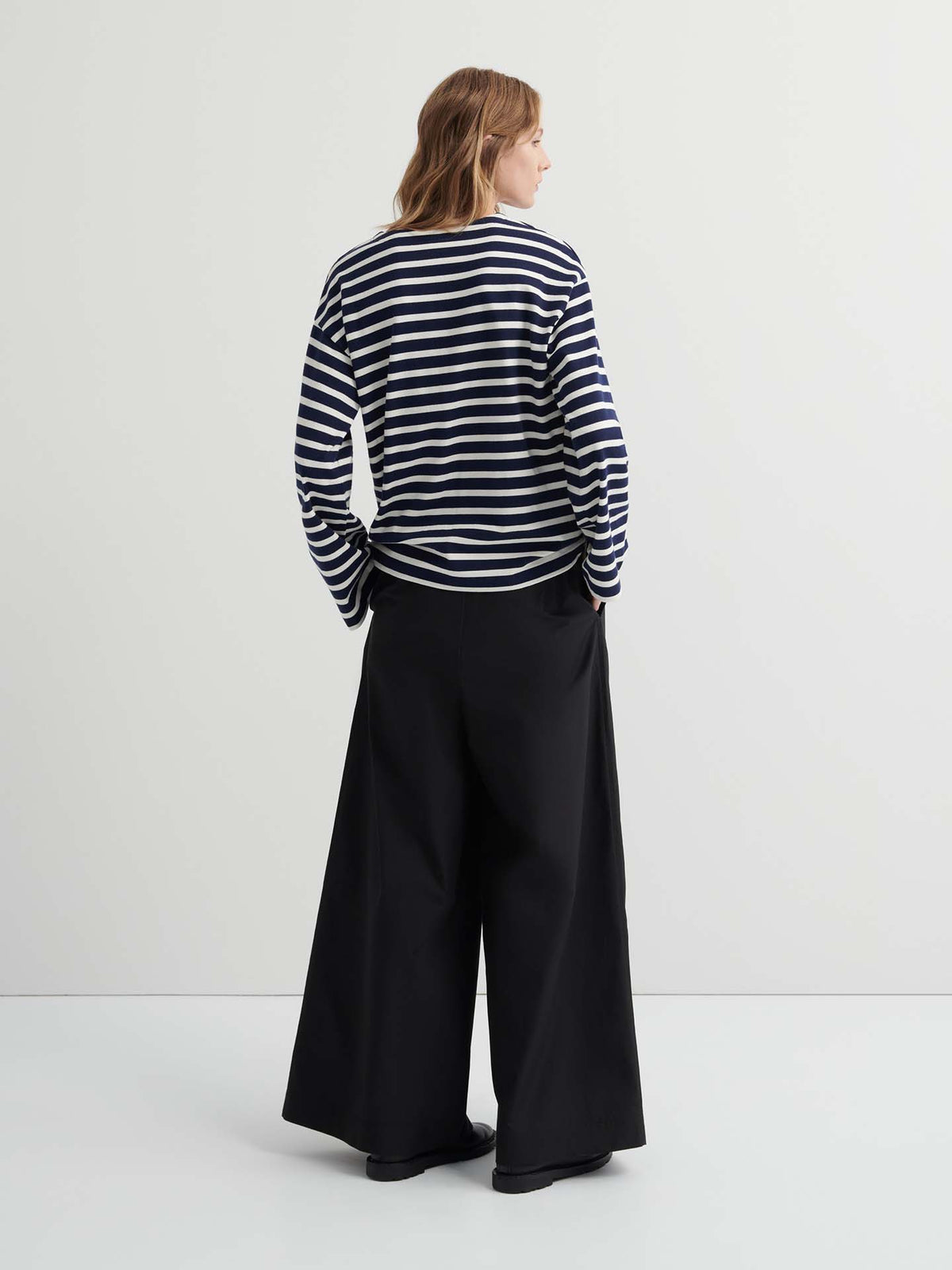 A person stands facing away, wearing the Pierre Tee – Uniform Stripe, a black and white striped oversized long-sleeve shirt, paired with wide-leg black pants against a plain, light-colored background.