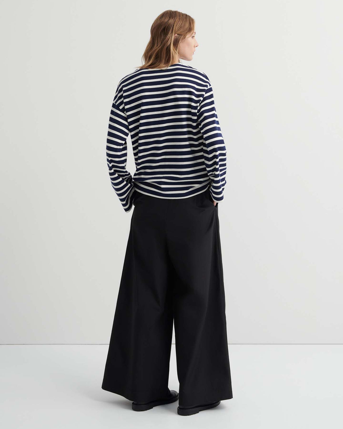 A person stands facing away, wearing the Pierre Tee – Uniform Stripe, a black and white striped oversized long-sleeve shirt, paired with wide-leg black pants against a plain, light-colored background.