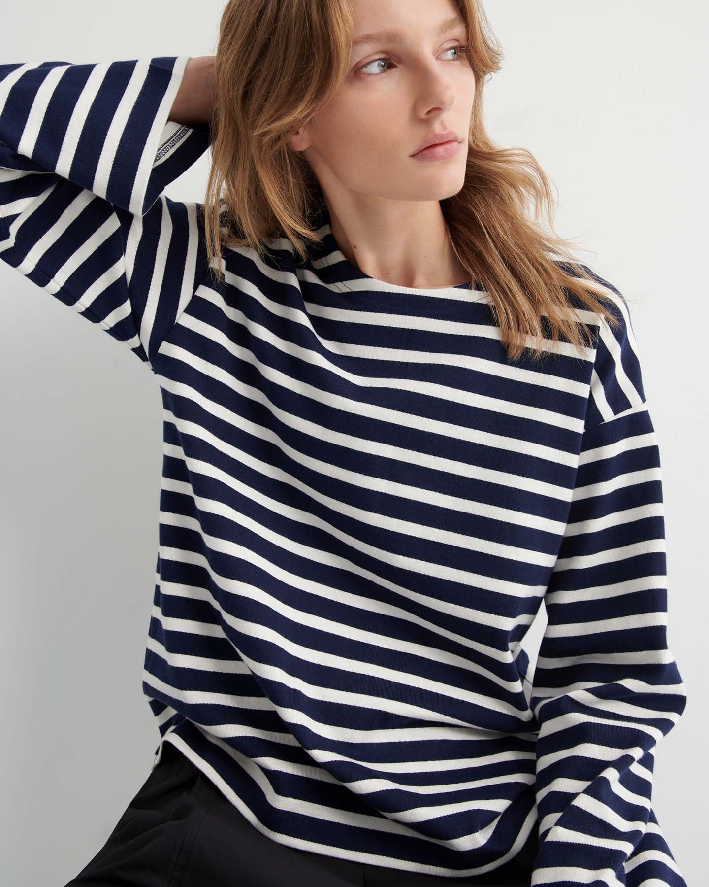 Wearing the Pierre Tee – Uniform Stripe, a relaxed-fit navy and white striped long-sleeve shirt, a person sits with one hand behind their head, gazing to the side.