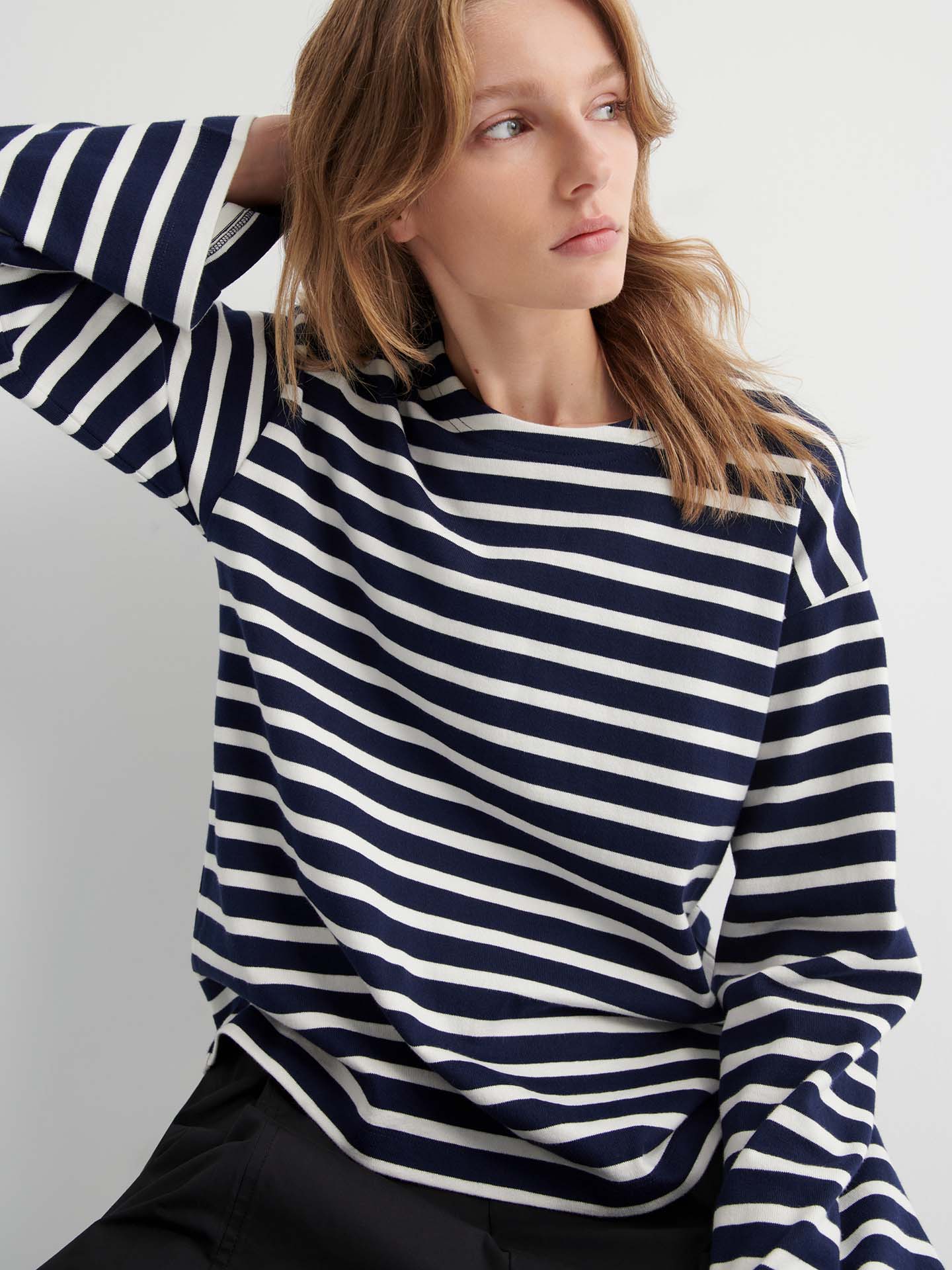 Wearing the Pierre Tee – Uniform Stripe, a relaxed-fit navy and white striped long-sleeve shirt, a person sits with one hand behind their head, gazing to the side.
