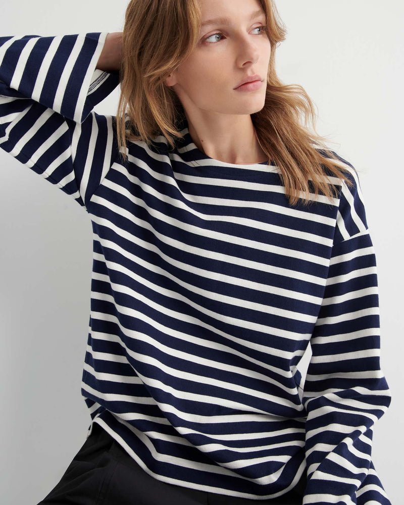 Wearing the Pierre Tee – Uniform Stripe, a relaxed-fit navy and white striped long-sleeve shirt, a person sits with one hand behind their head, gazing to the side.