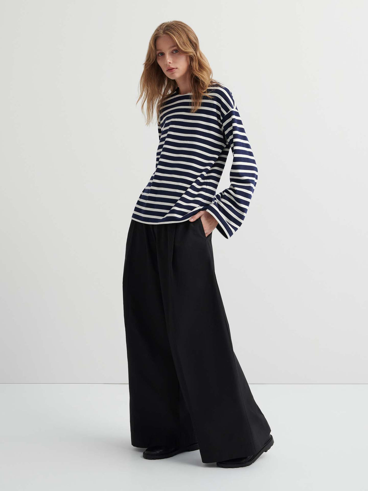 A person stands against a plain background, wearing the Pierre Tee – Uniform Stripe (a black and white oversized long-sleeve shirt) with wide-leg black pants, hands in pockets.