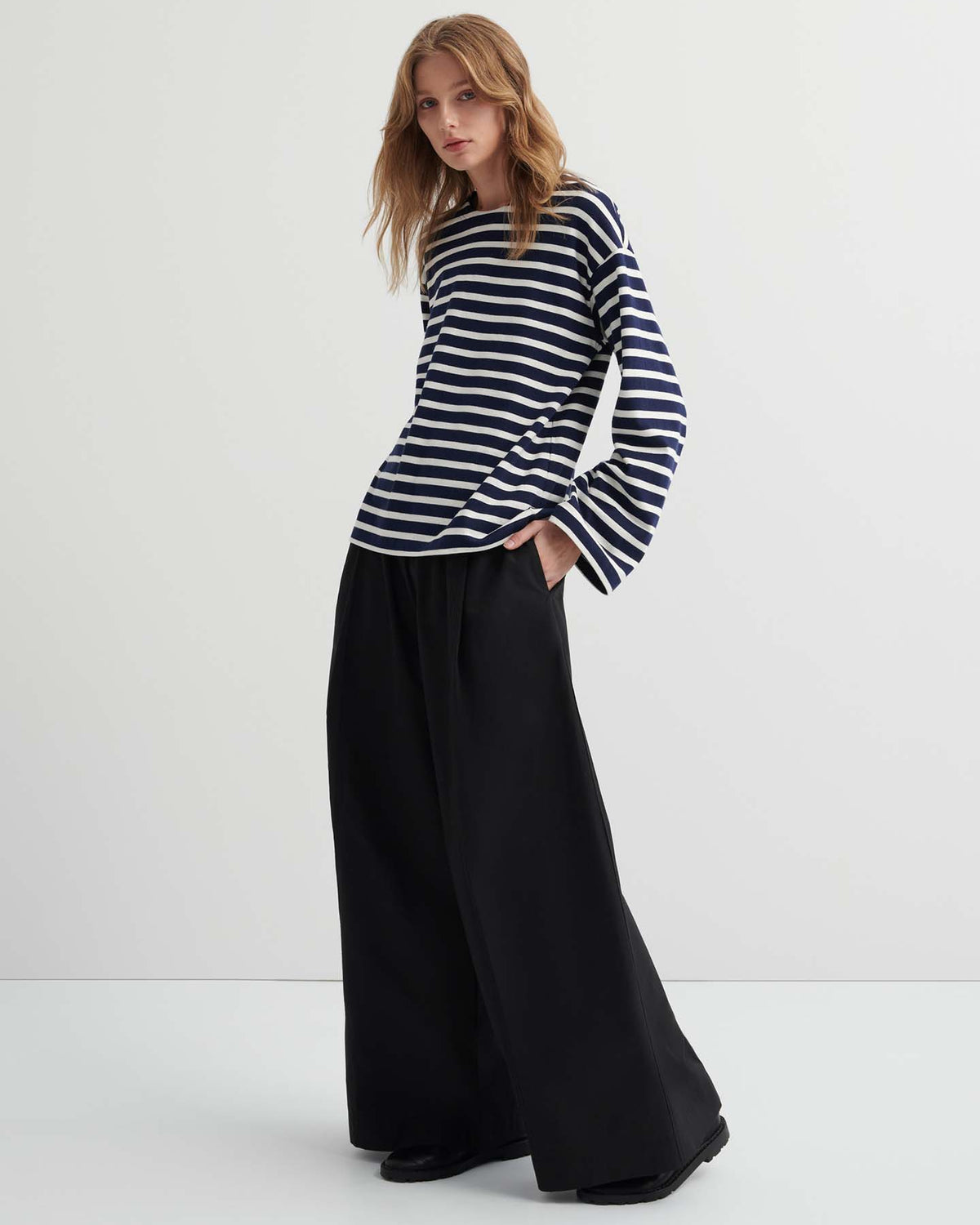 A person stands against a plain background, wearing the Pierre Tee – Uniform Stripe (a black and white oversized long-sleeve shirt) with wide-leg black pants, hands in pockets.