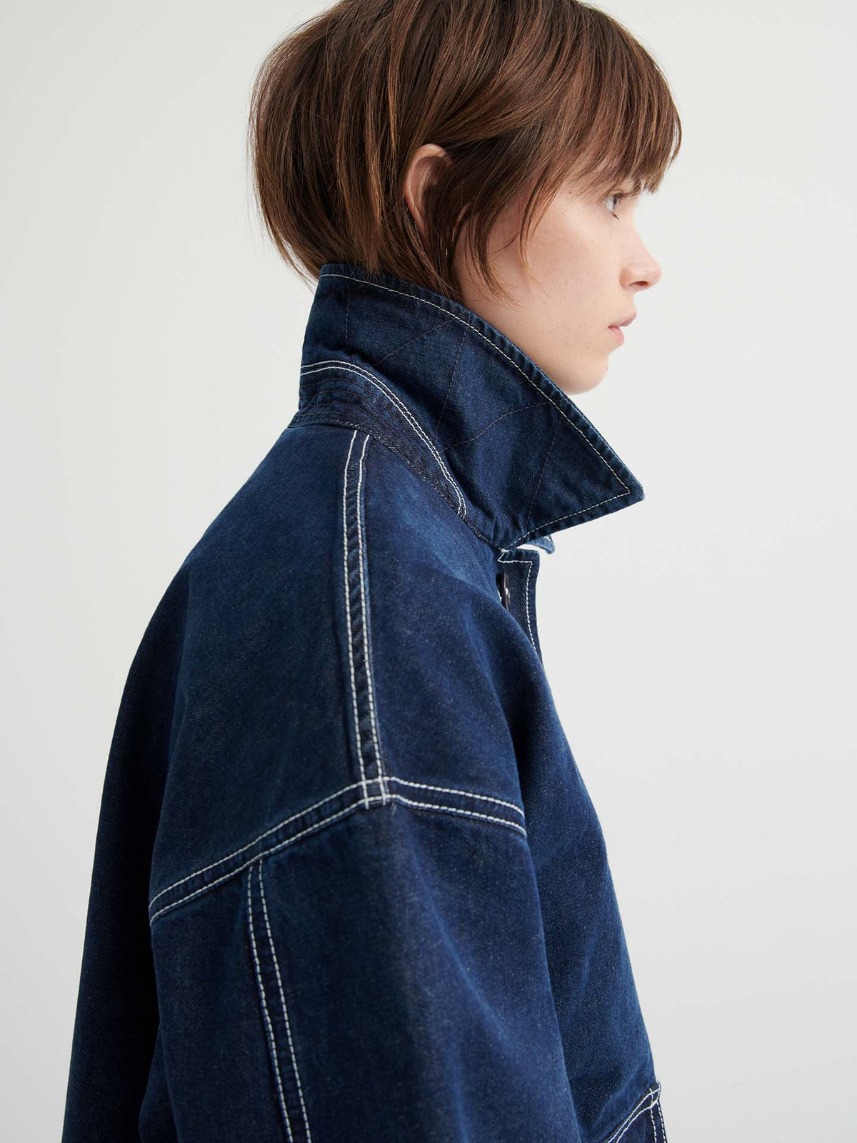 A person with short brown hair in profile wears the Portrait Jacket – Indigo, a dark blue, boxy-fit jacket featuring prominent white stitching and an upturned collar, set against a plain light background.