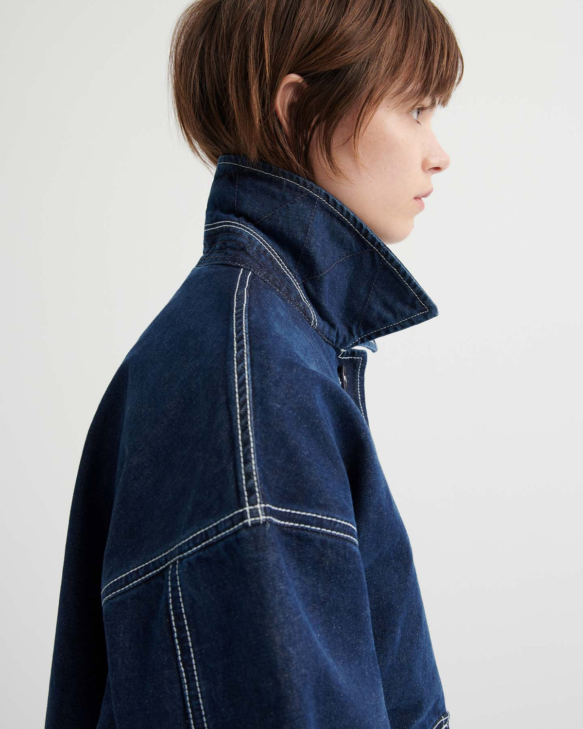 A person with short brown hair in profile wears the Portrait Jacket – Indigo, a dark blue, boxy-fit jacket featuring prominent white stitching and an upturned collar, set against a plain light background.