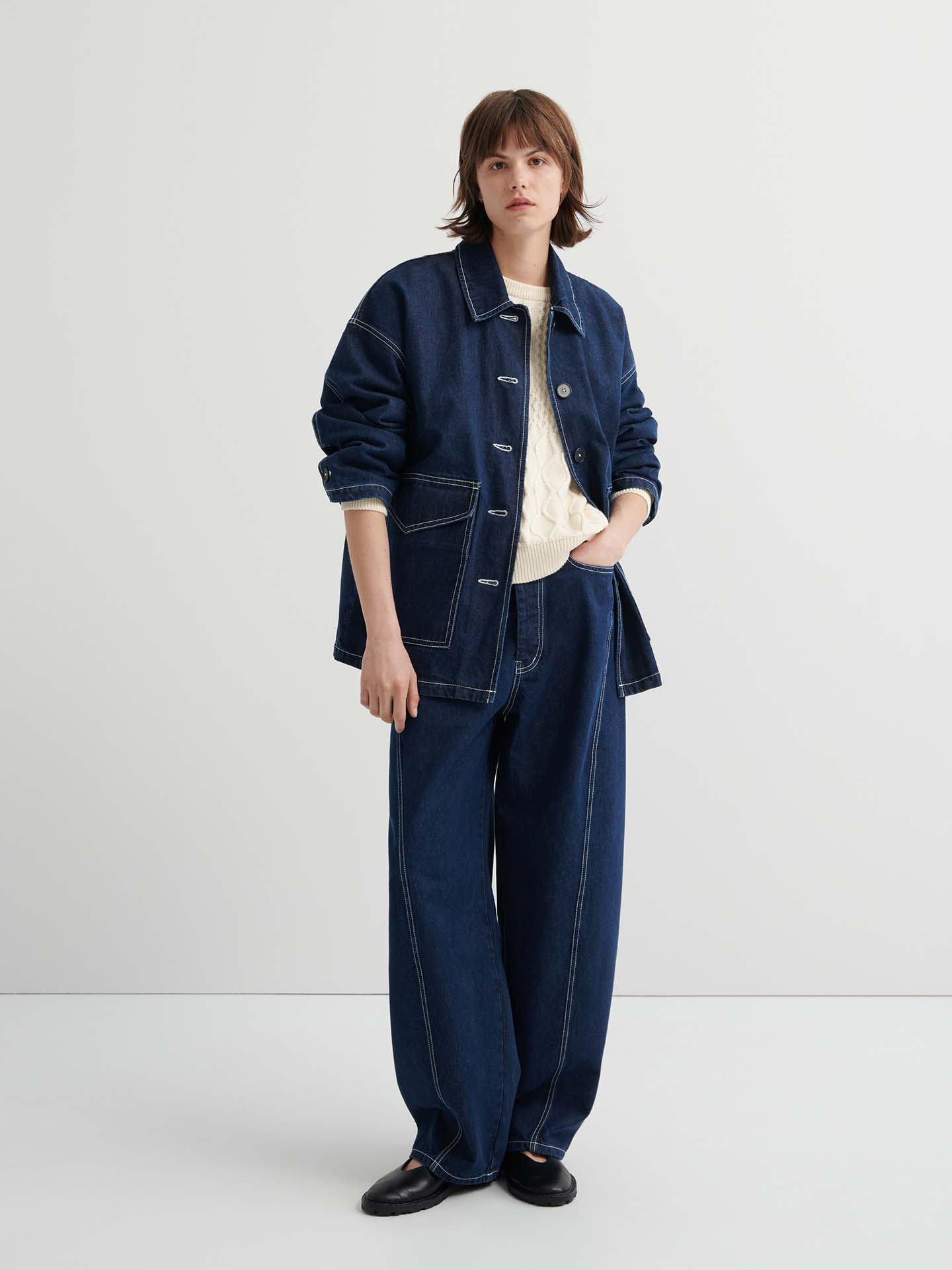 A person stands against a plain background wearing the Portrait Jacket – Indigo with matching wide-leg jeans over a cream sweater, completed with black shoes.