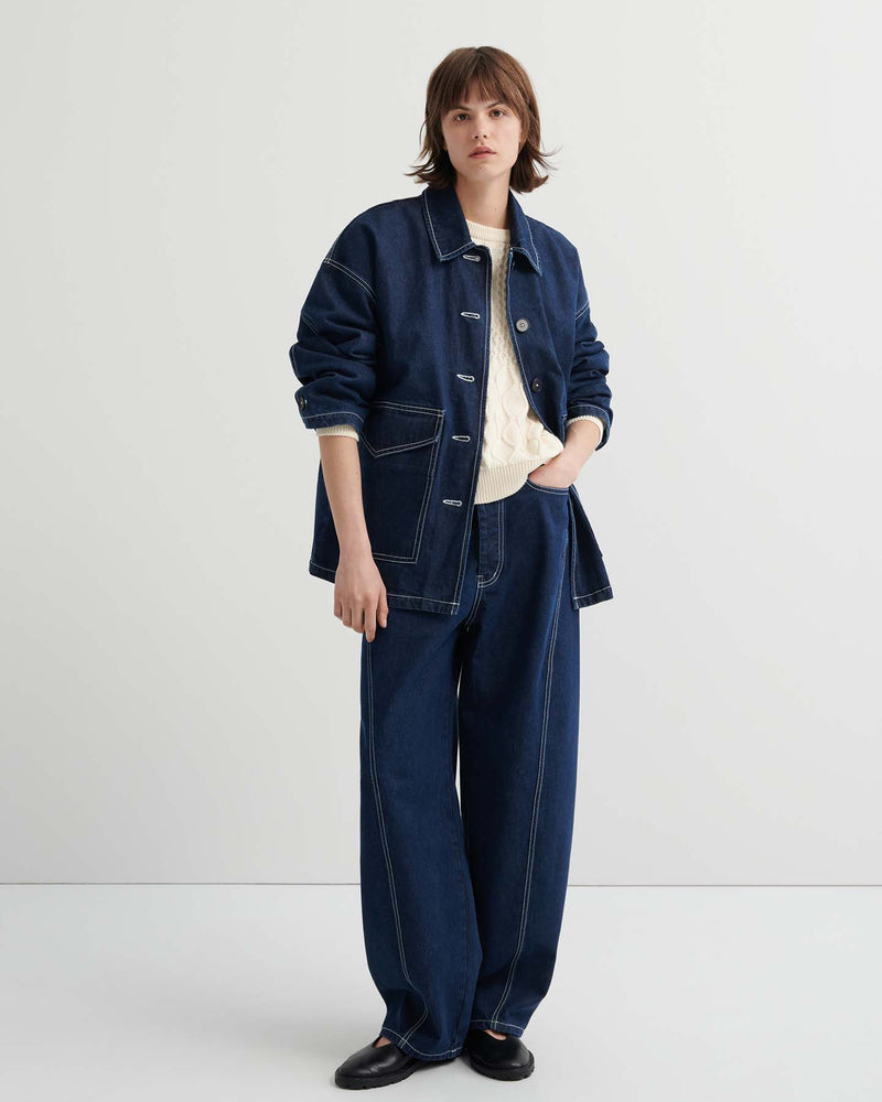 A person stands against a plain background wearing the Portrait Jacket – Indigo with matching wide-leg jeans over a cream sweater, completed with black shoes.