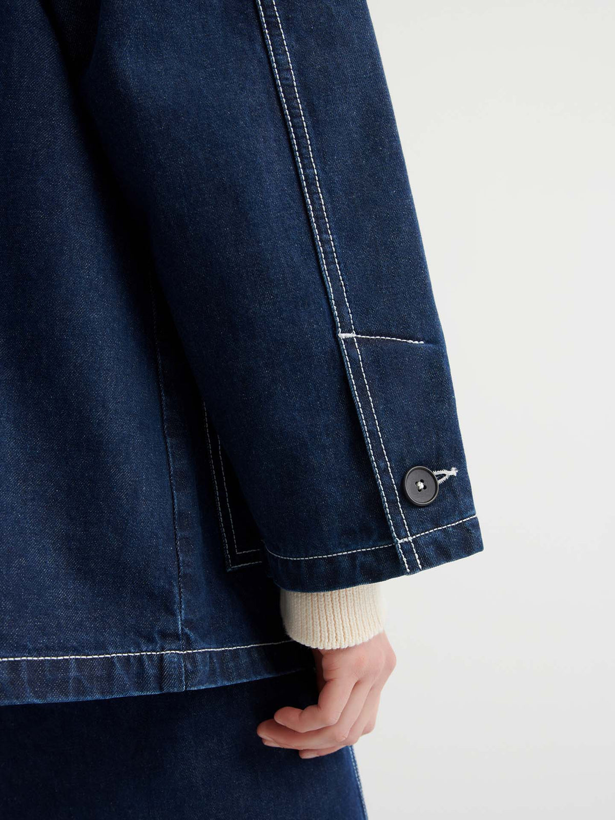 A person wears the Portrait Jacket – Indigo, a dark, boxy fit style with matching pants and white contrast stitching, featuring a close-up of the sleeve, cuff button, and part of their hand.