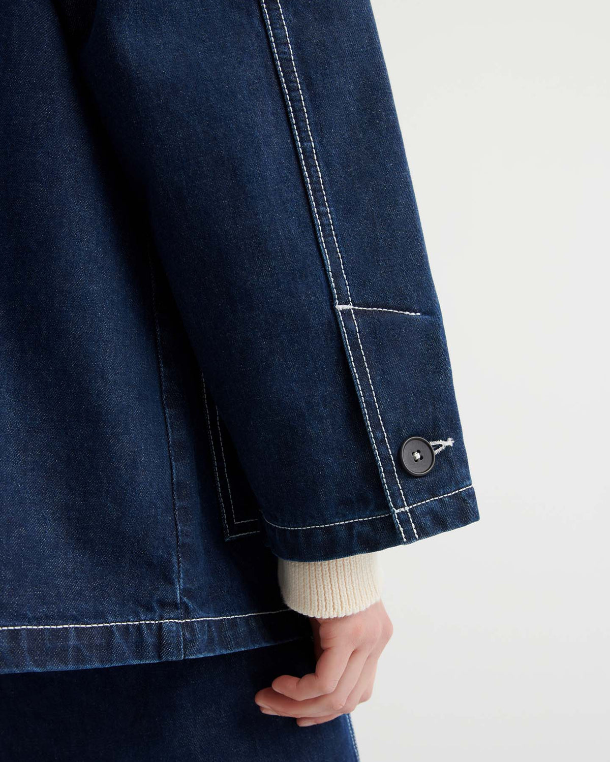 A person wears the Portrait Jacket – Indigo, a dark, boxy fit style with matching pants and white contrast stitching, featuring a close-up of the sleeve, cuff button, and part of their hand.